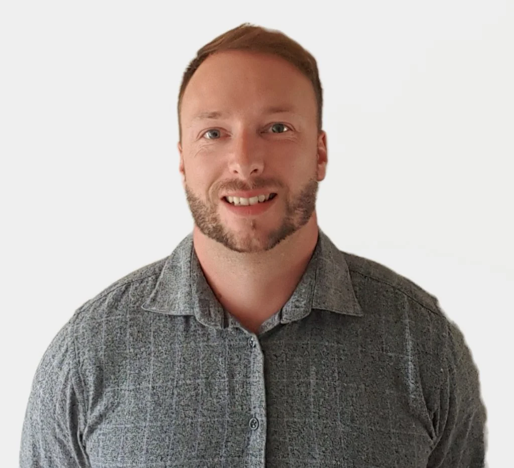 A man with short hair and a beard, smiling, wearing a gray patterned button-up shirt, against a plain white background.