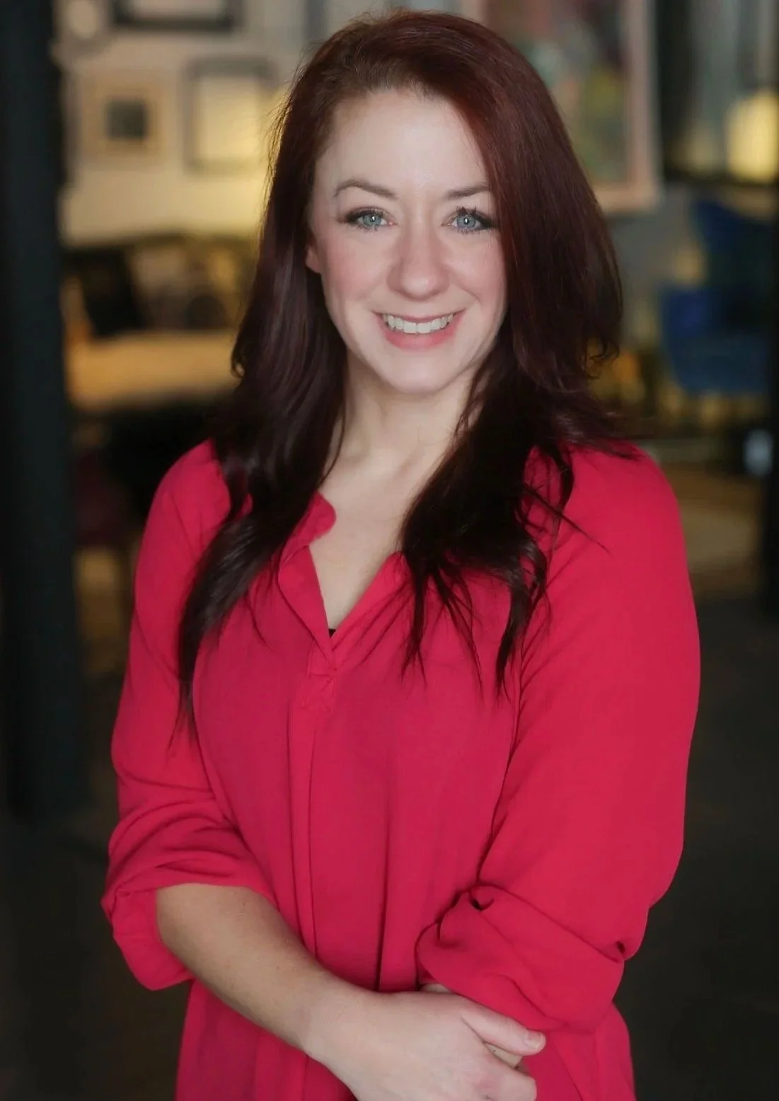 A woman with long, wavy red hair and blue eyes smiling, wearing a pink blouse, standing indoors with a blurred background.