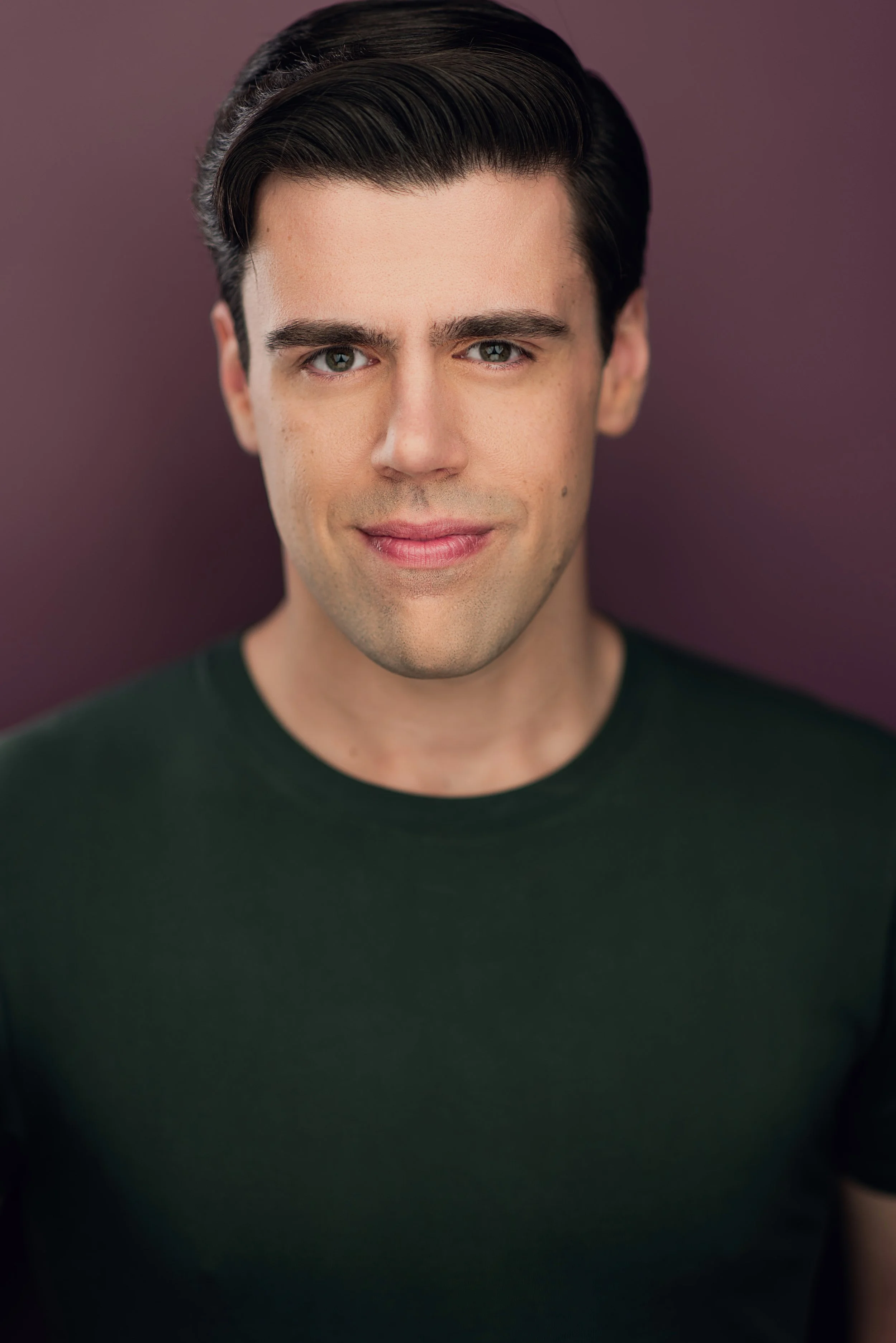 Close-up portrait of a young man with dark hair styled back, fair skin, blue eyes, and a slight smile, wearing a black shirt against a purple background.