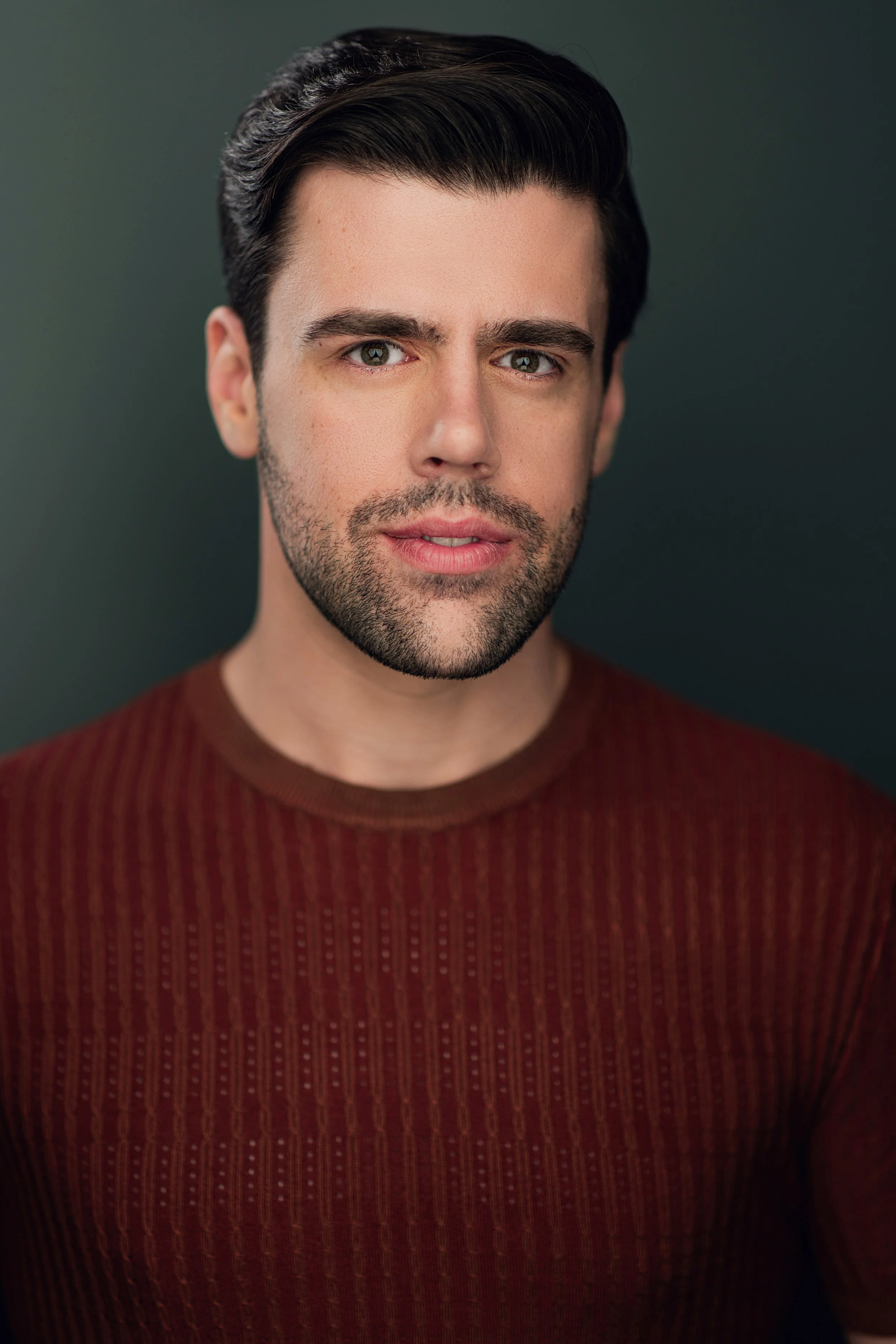 Close-up portrait of a man with dark hair, hazel eyes, and a light beard, wearing a burnt orange sweater, against a dark background.