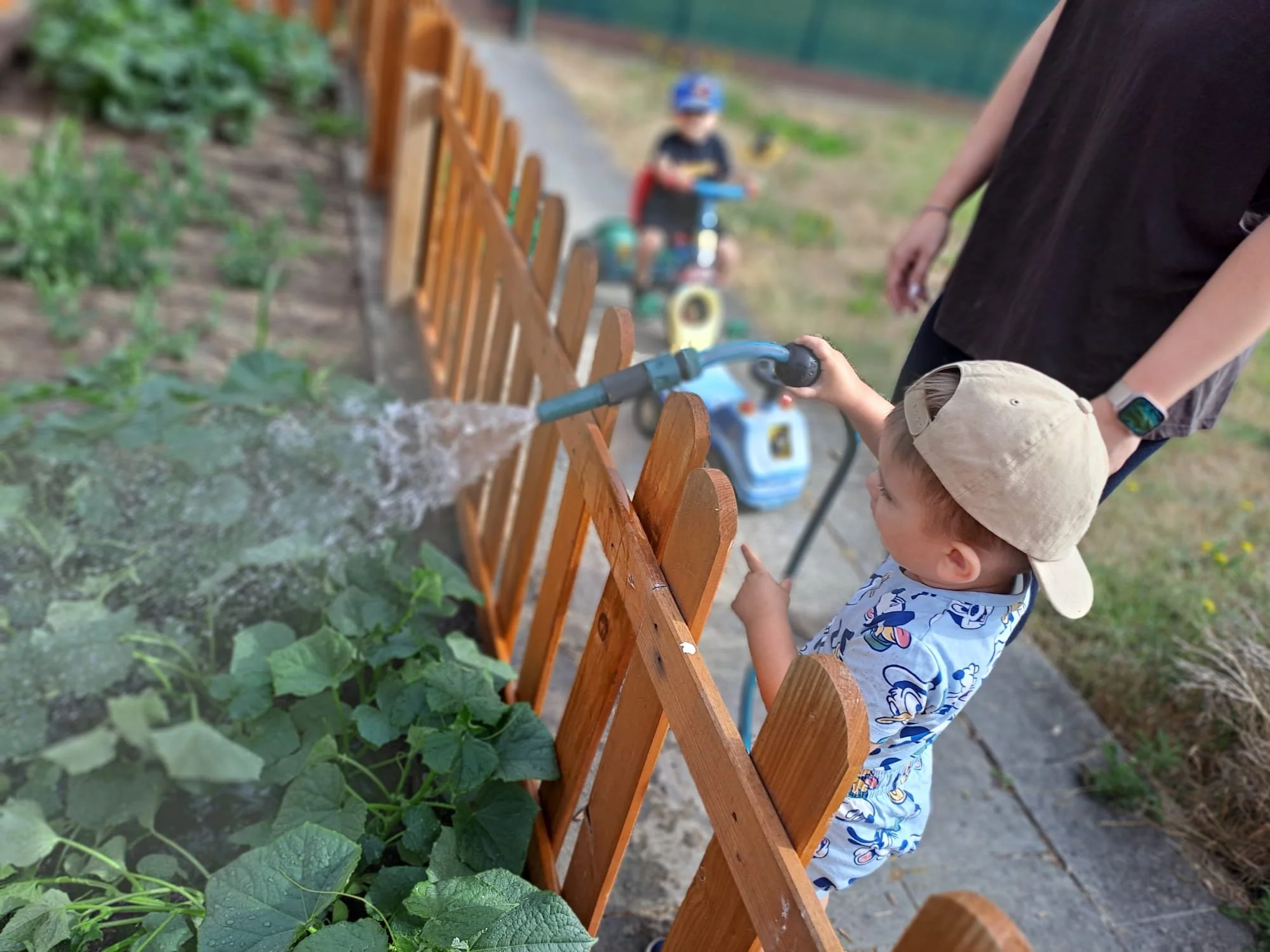Deux jeunes enfants regardent de près des plantes dans un jardin, avec un adulte qui tient la poignée d'un petit vélo électrique en arrière-plan, et un petit enfant en casquette beige devant eux.