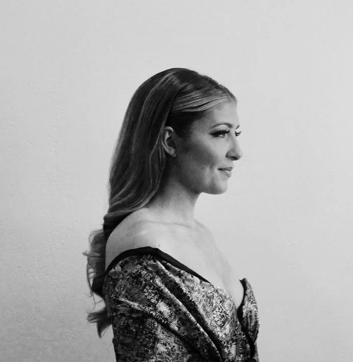 A black and white side profile of a woman with long hair, wearing an off-the-shoulder top, standing against a plain wall.