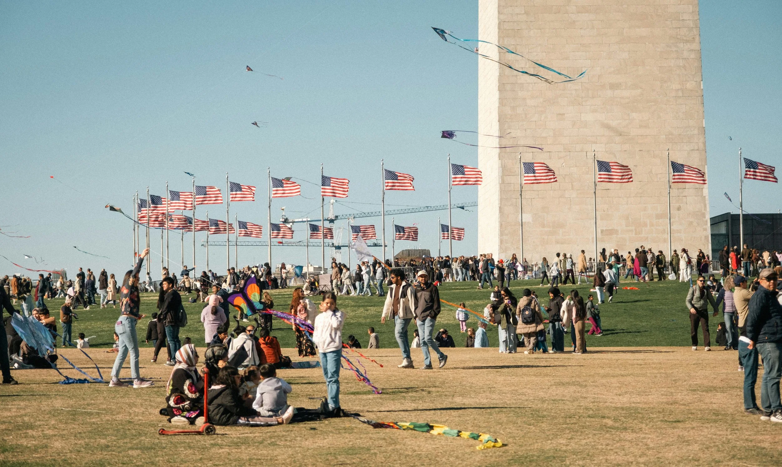 People flying kites on the National Mall in Washington, D.C. with the Washington Monument in the background, American flags lining the area, and various groups and individuals enjoying the outdoor event.