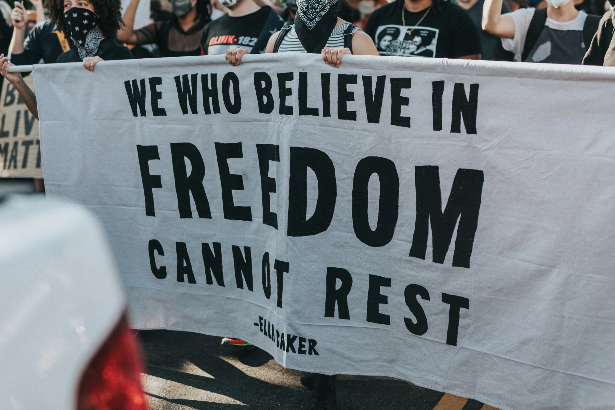 People participating in a protest march, holding a large white banner with the message 'WE WHO BELIEVE IN FREEDOM CANNOT BE BUS' written in bold black letters, surrounded by other protesters.