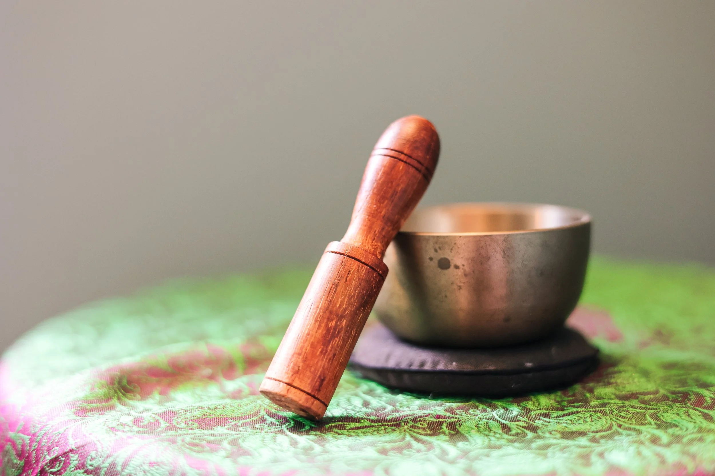 Close-up of a wooden striker resting against a metal Tibetan singing bowl, with a soft green and pink cloth underneath.
