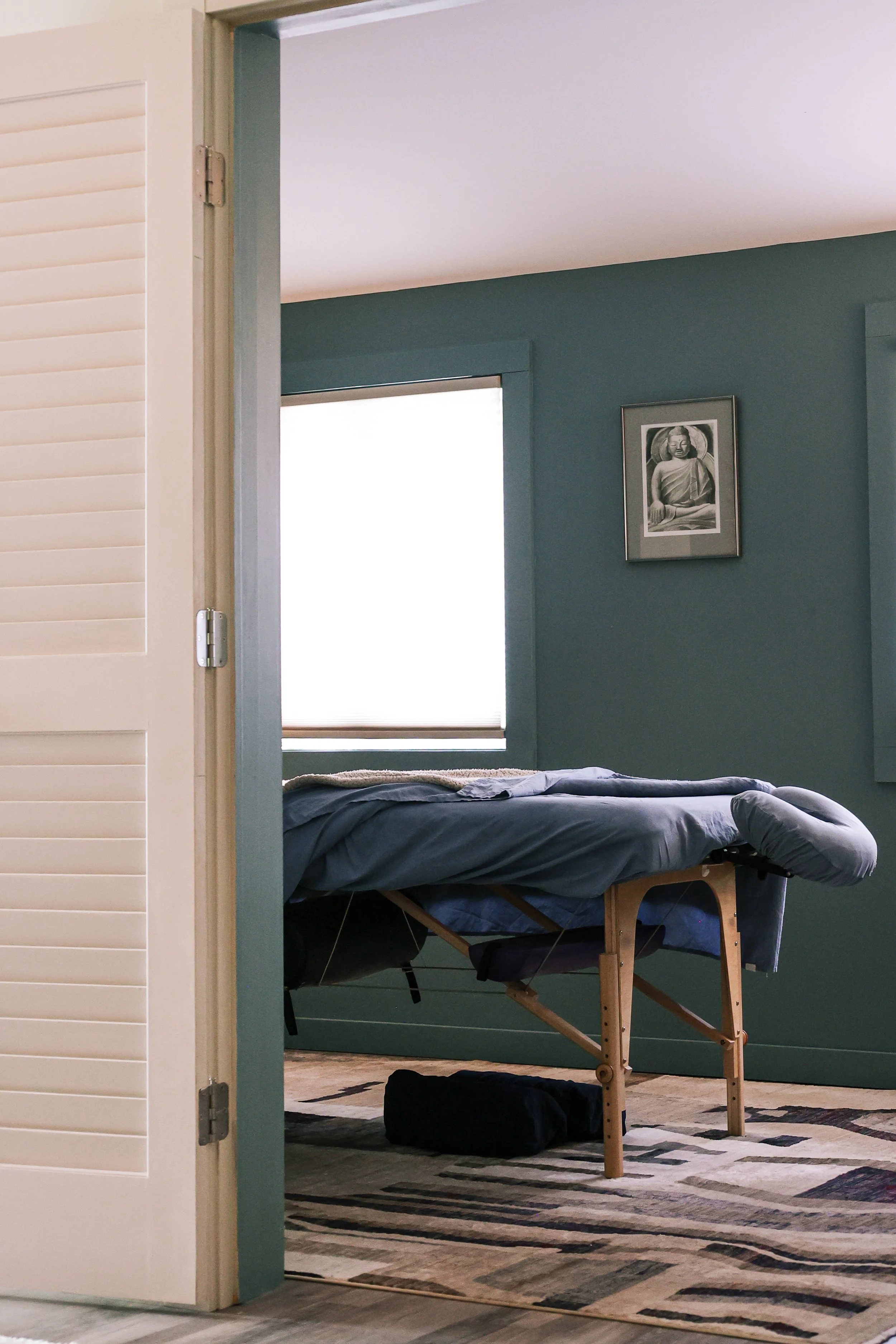 A massage table set up in a room with dark teal walls and a framed black-and-white picture of Buddha on the wall. There is a window with white blinds and a patterned rug on the floor.