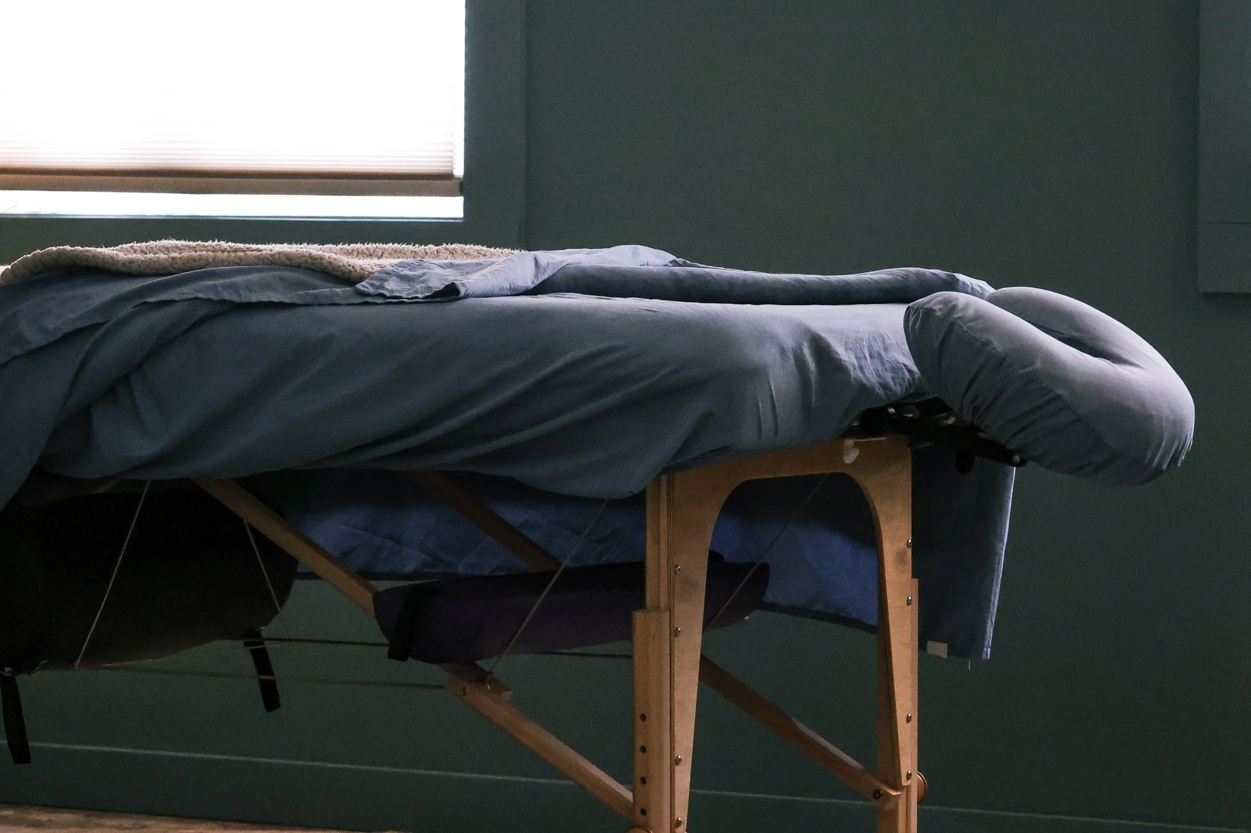 A bed with blue sheets and a beige blanket, placed on a wooden frame in a bedroom with green walls and a window with blinds.