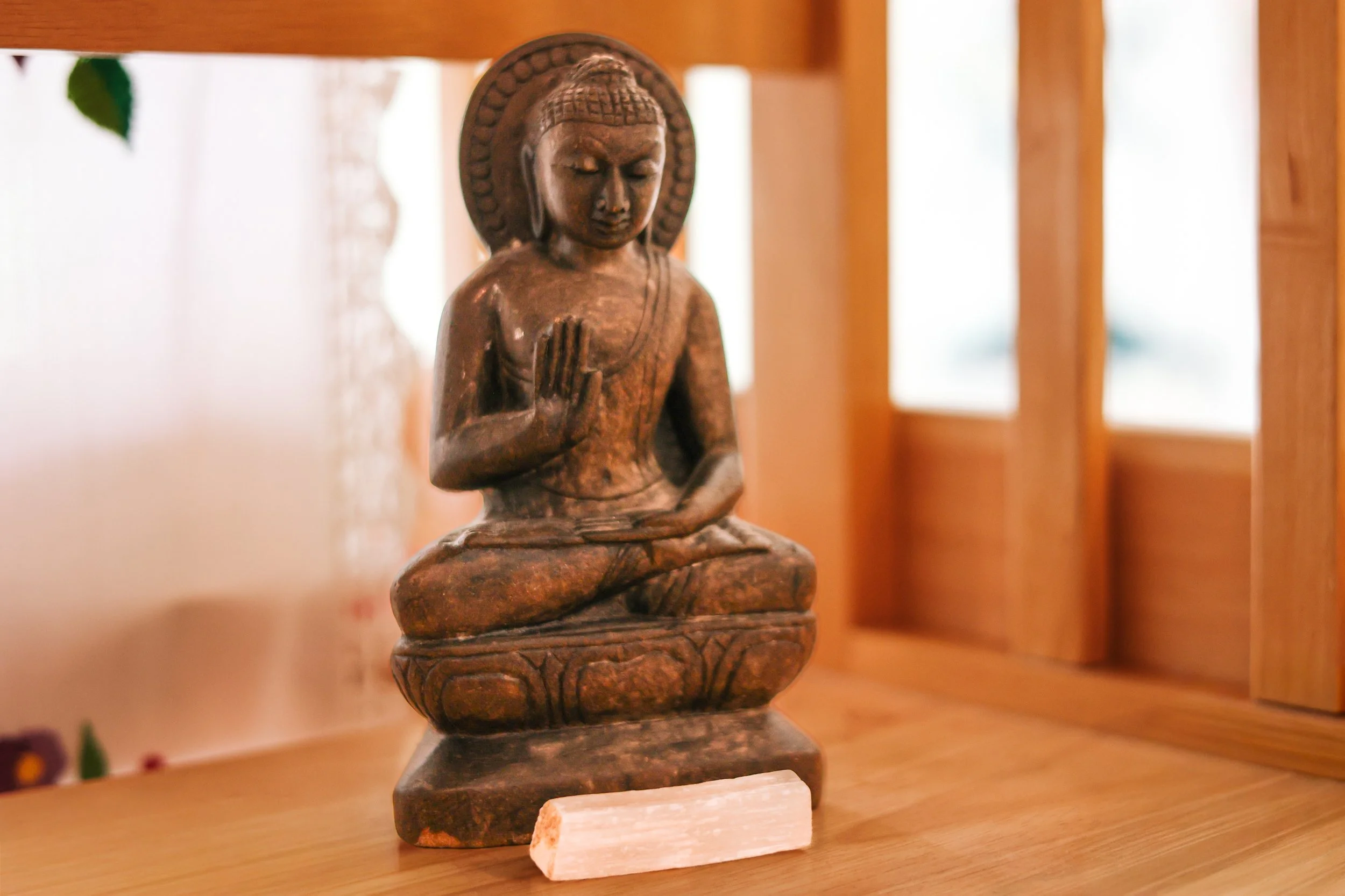 Wooden sculpture of a seated meditative figure with a halo-like circle behind the head, placed on a small platform, in a room with wooden furniture and windows in the background.