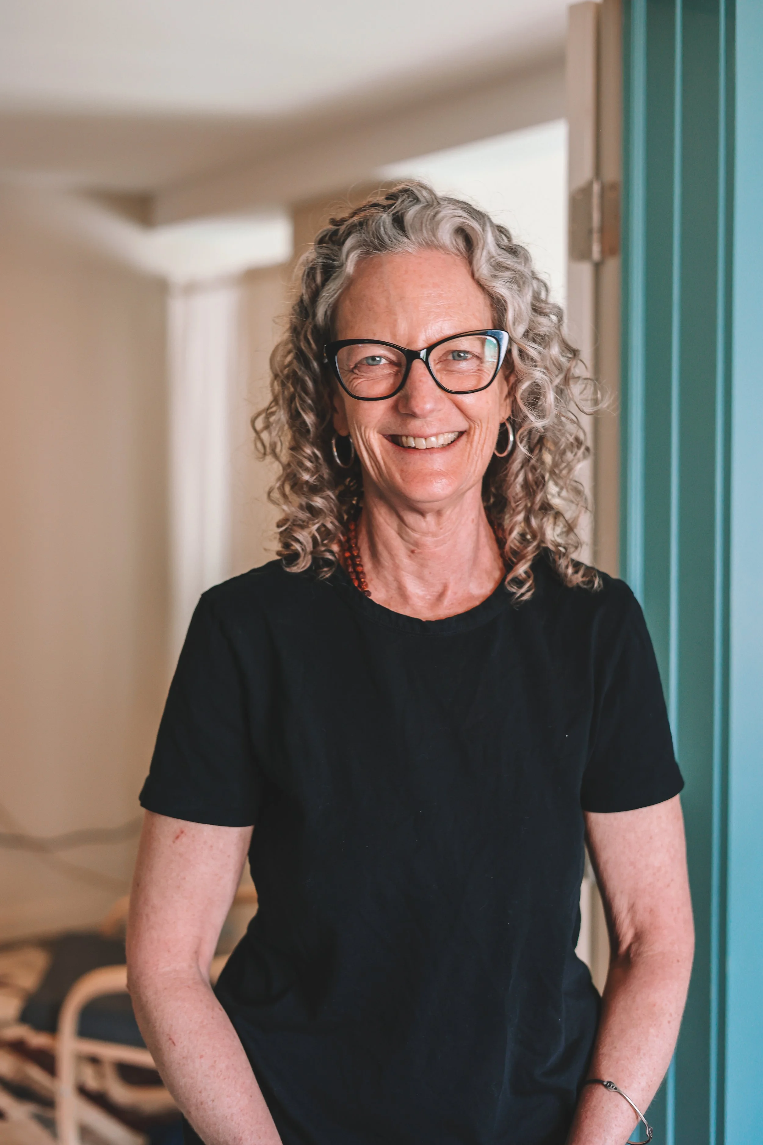 A mature woman with curly gray hair, glasses, and hoop earrings, smiling while standing indoors in front of a door and beige wall.