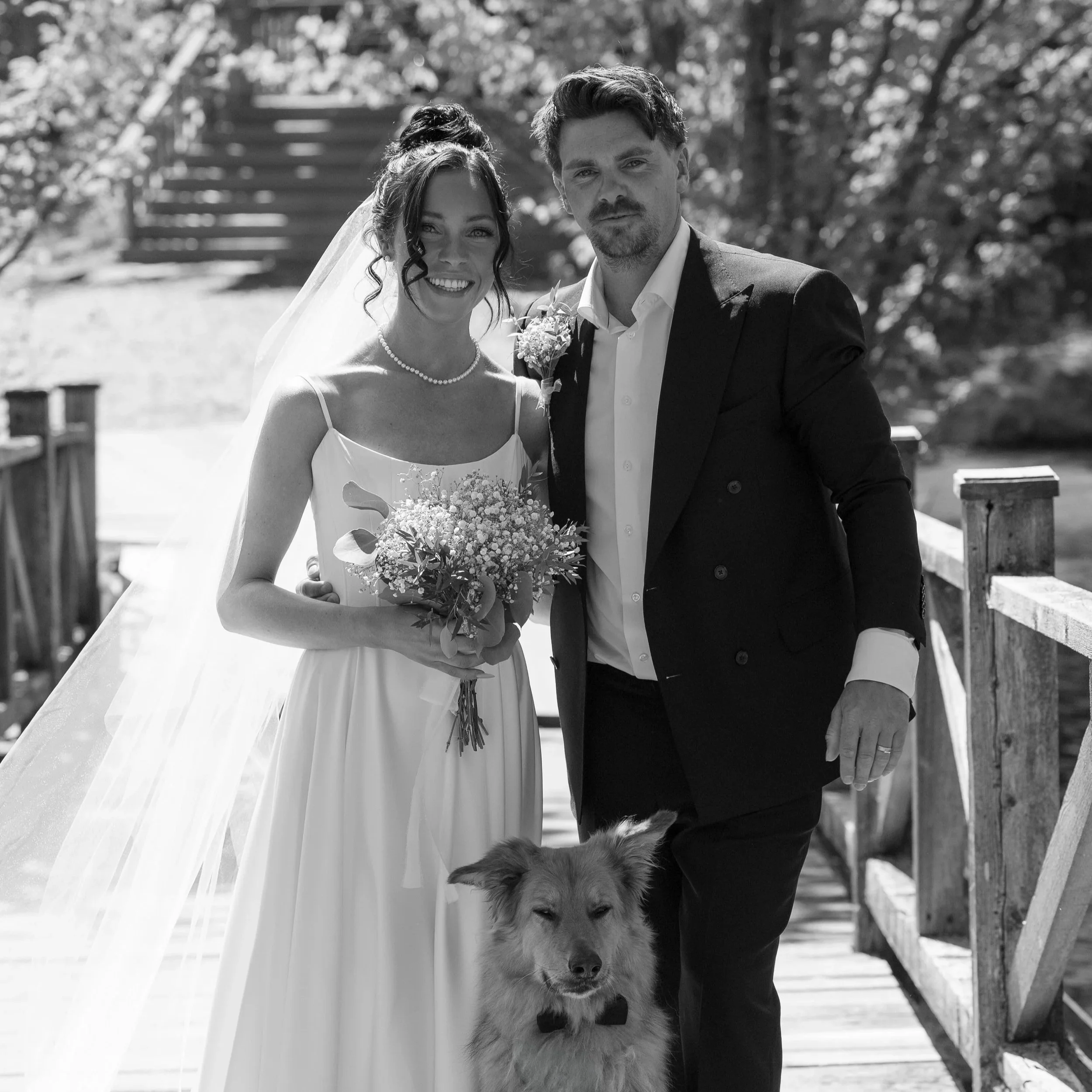 Black and white photo of a bride and groom with a dog at their wedding. The bride is smiling and holding a bouquet, while the groom is standing beside her with a serious expression. The dog is sitting in front of them.