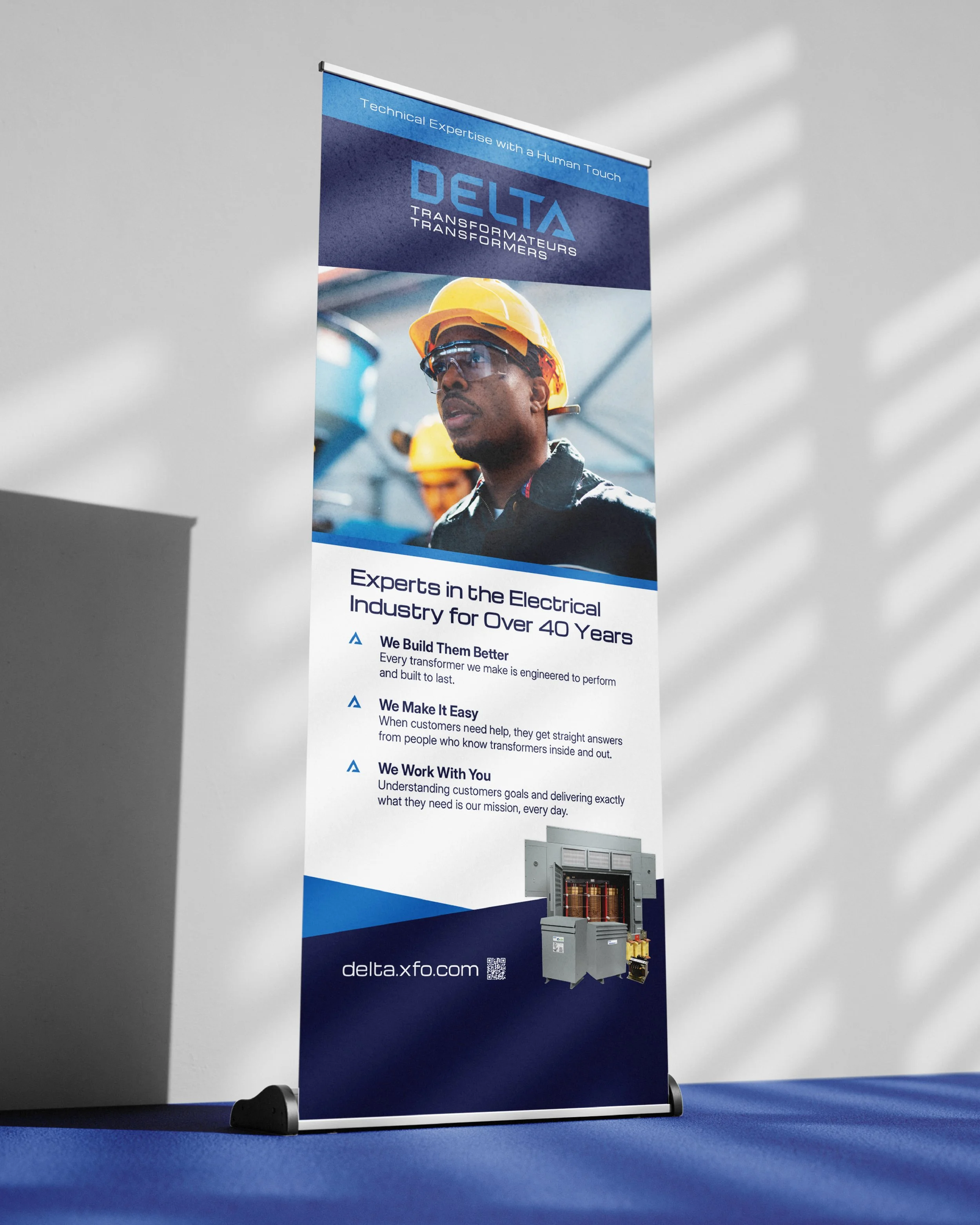 A tall, vertical promotional banner for Delta Transformers on a blue carpeted floor with a plain gray wall background. The banner features an image of a person wearing a yellow safety helmet, glasses, and a dark work uniform. The top section has the text "Technical Expertise with a Human Touch," the middle displays the Delta Transformers logo and slogan, and the lower section lists the company's key services and website, delta.xfo.com, alongside a QR code.