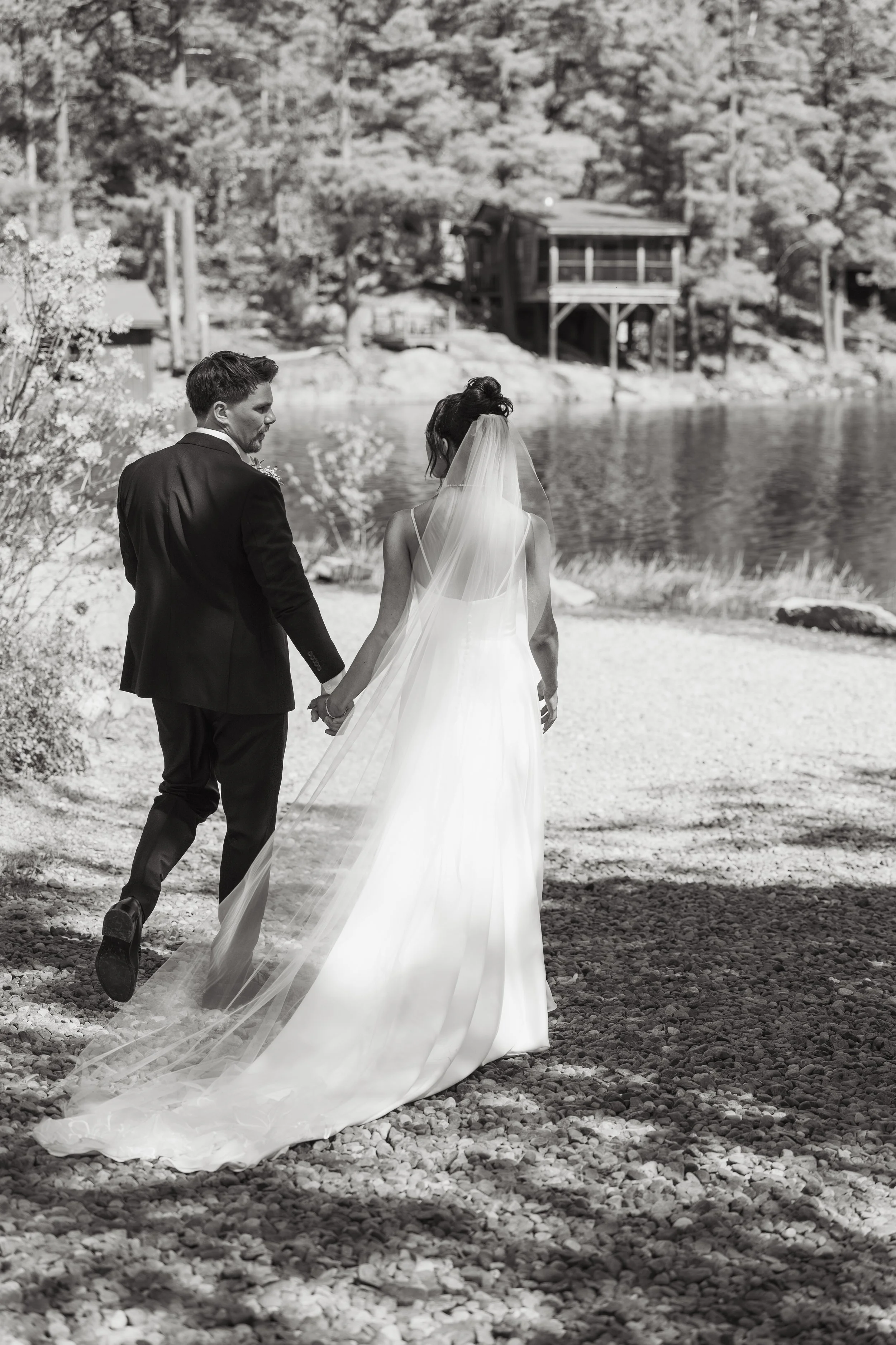 Black and white photo of a bride and groom holding hands walking on rocky ground near a body of water, with trees and a house on stilts in the background.