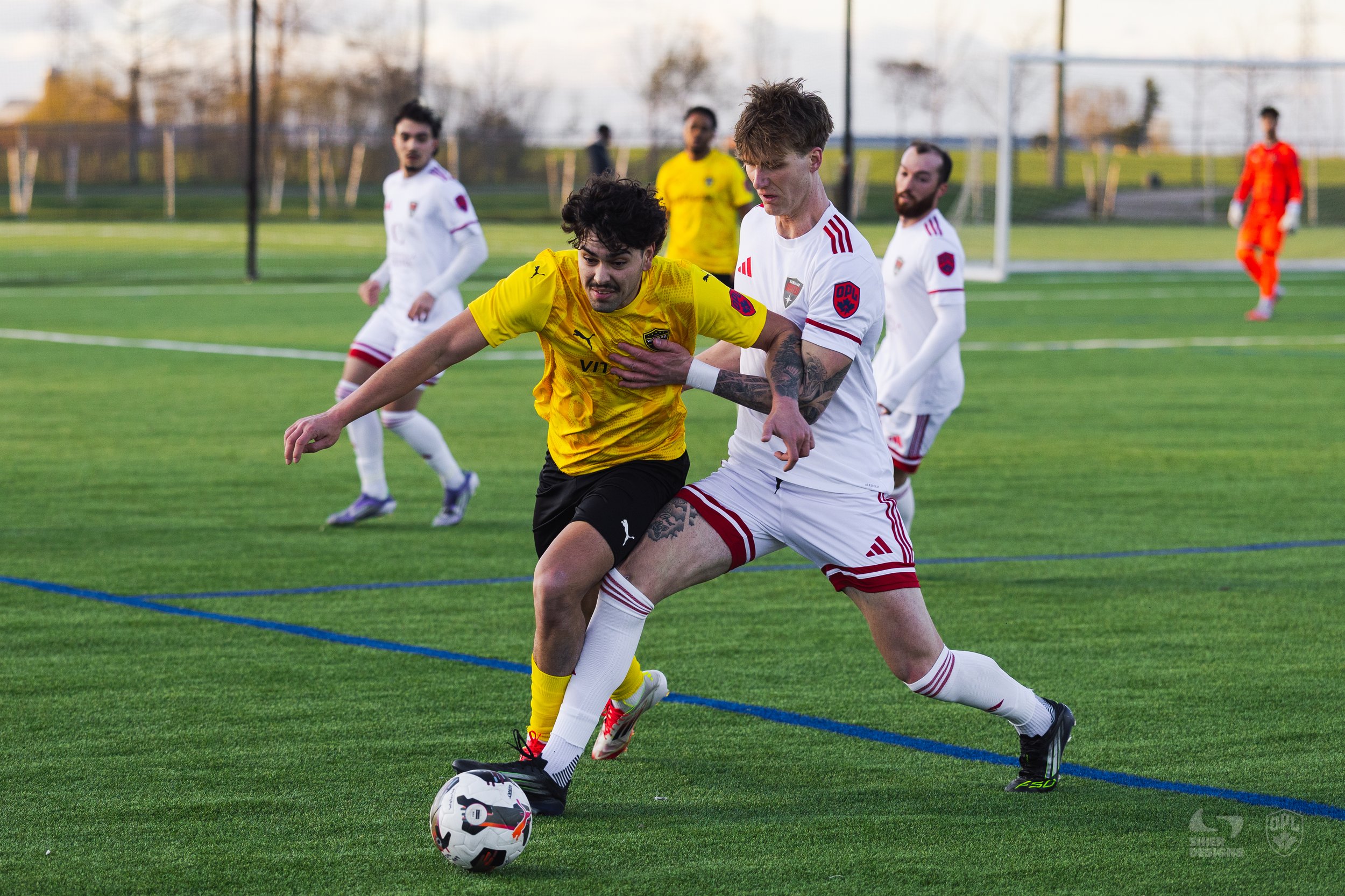 Two soccer players, one in a yellow jersey and the other in a white jersey, compete for the ball on a green field. The player in yellow is leaning forward, while the player in white has his arm on the opponent and is positioned to kick the ball. In the background, other players and a goal are visible on a soccer field.