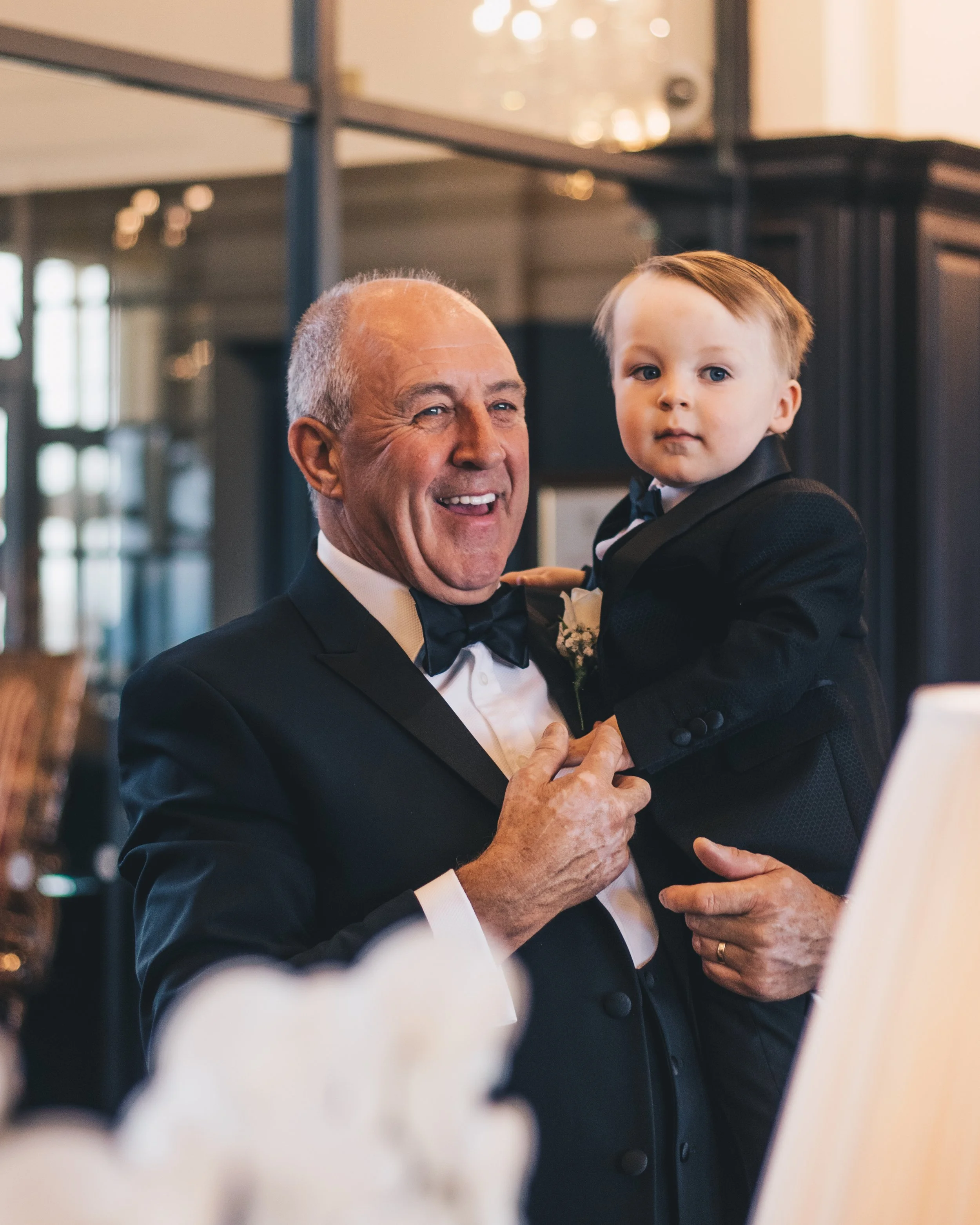 An elderly man in a tuxedo is smiling and holding a young boy in a tuxedo, possibly at a formal event or wedding.