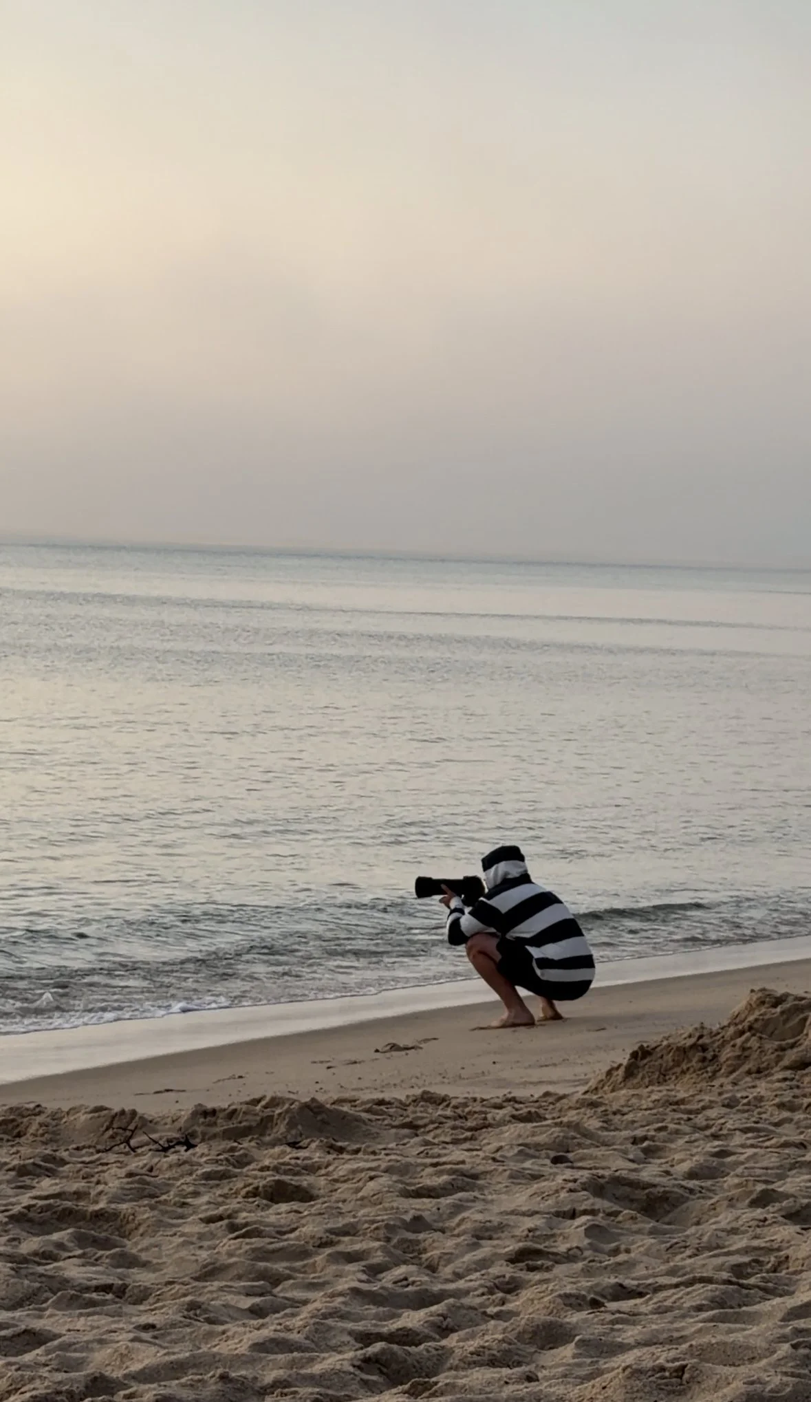 Person crouching on sandy beach near ocean, taking a photo with a camera, wearing a striped black and white long-sleeve shirt and shorts.