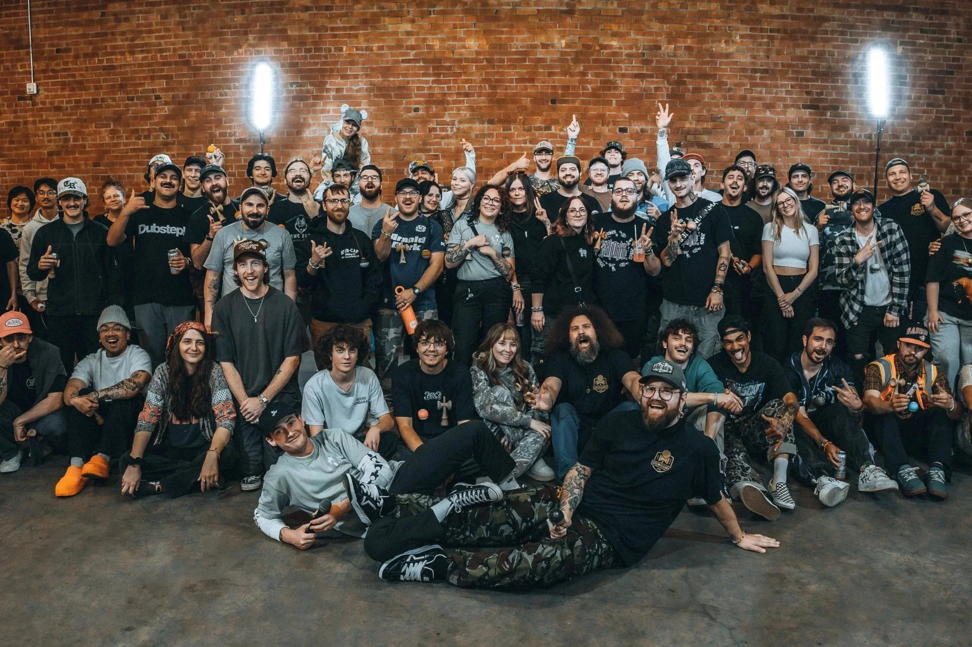 A diverse group of people posing together in front of a brick wall, some standing and some sitting, celebrating and smiling at an event, with two bright lights on stands behind them.