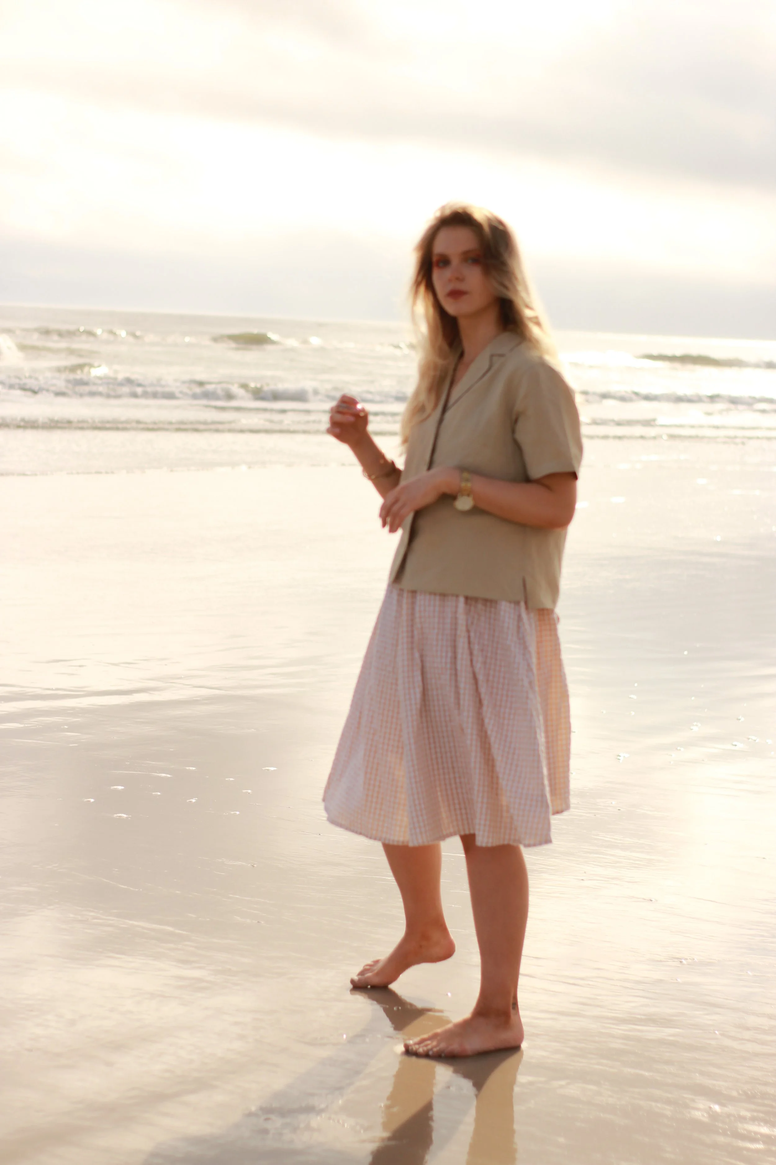 A woman with long hair wearing a beige shirt and pink checkered skirt standing in shallow water at the beach, with waves and a cloudy sky in the background.
