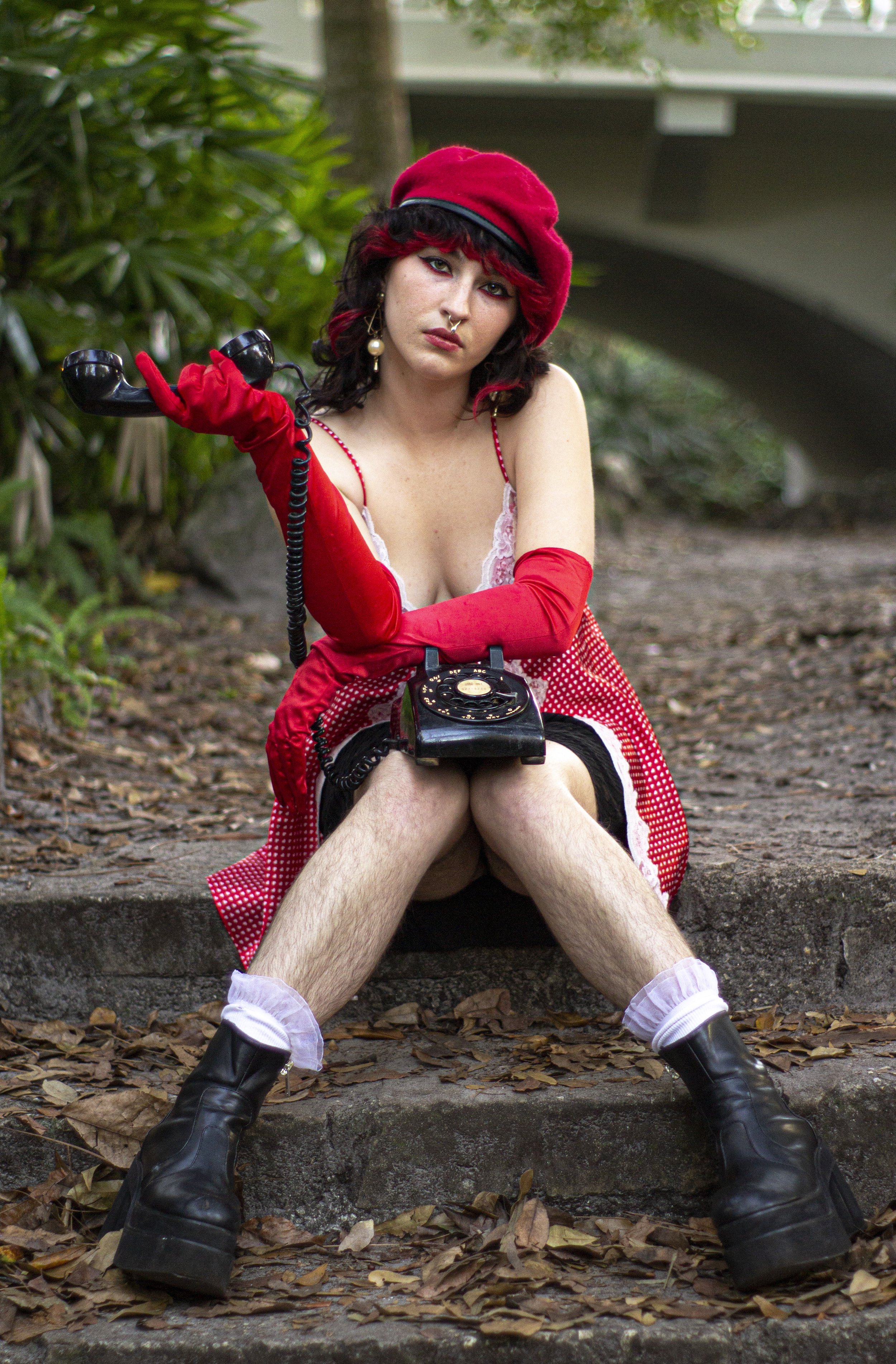 A woman wearing a red beret, red gloves, and a red checkered dress is sitting on outdoor steps with her legs apart. She holds a vintage black rotary phone, with an intense expression. The setting includes green foliage and a bridge in the background.