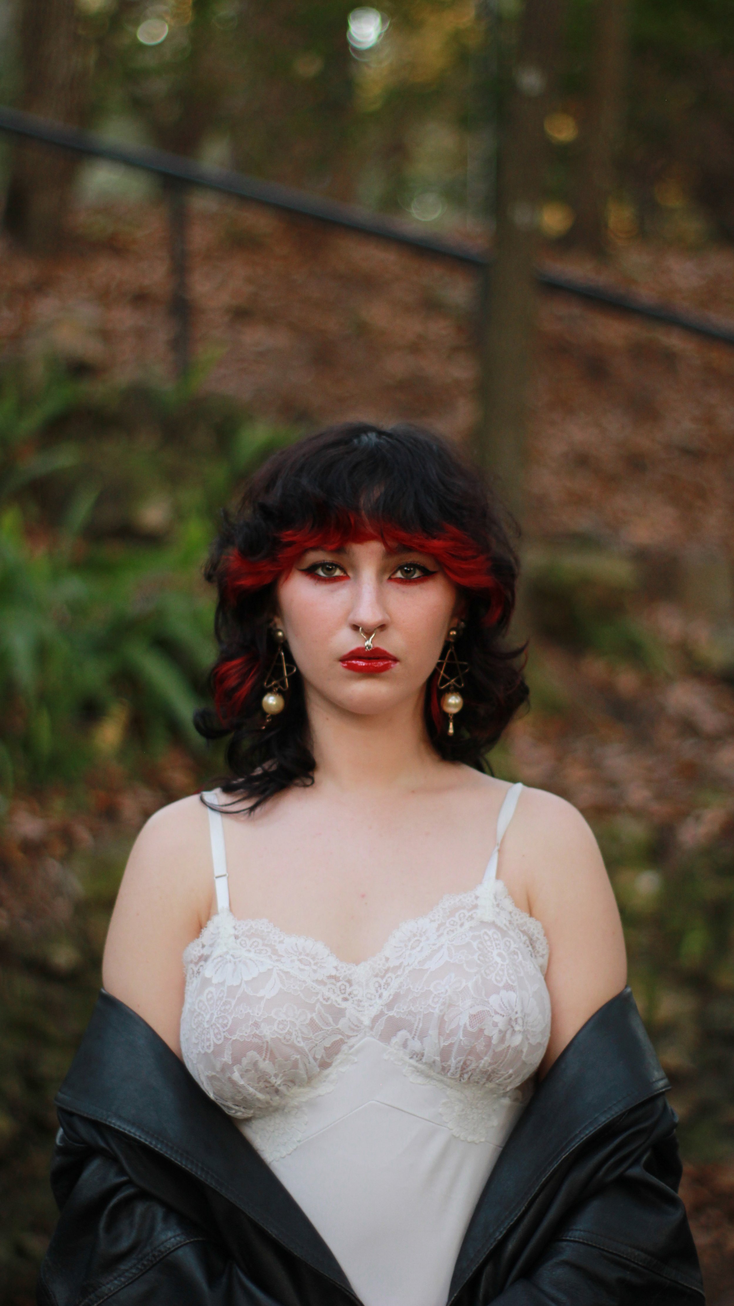 A woman with black and red hair, tan skin, wearing large earrings, a septum piercing, a lace white top, and a black jacket draped around her shoulders, standing outdoors with a blurred natural background.
