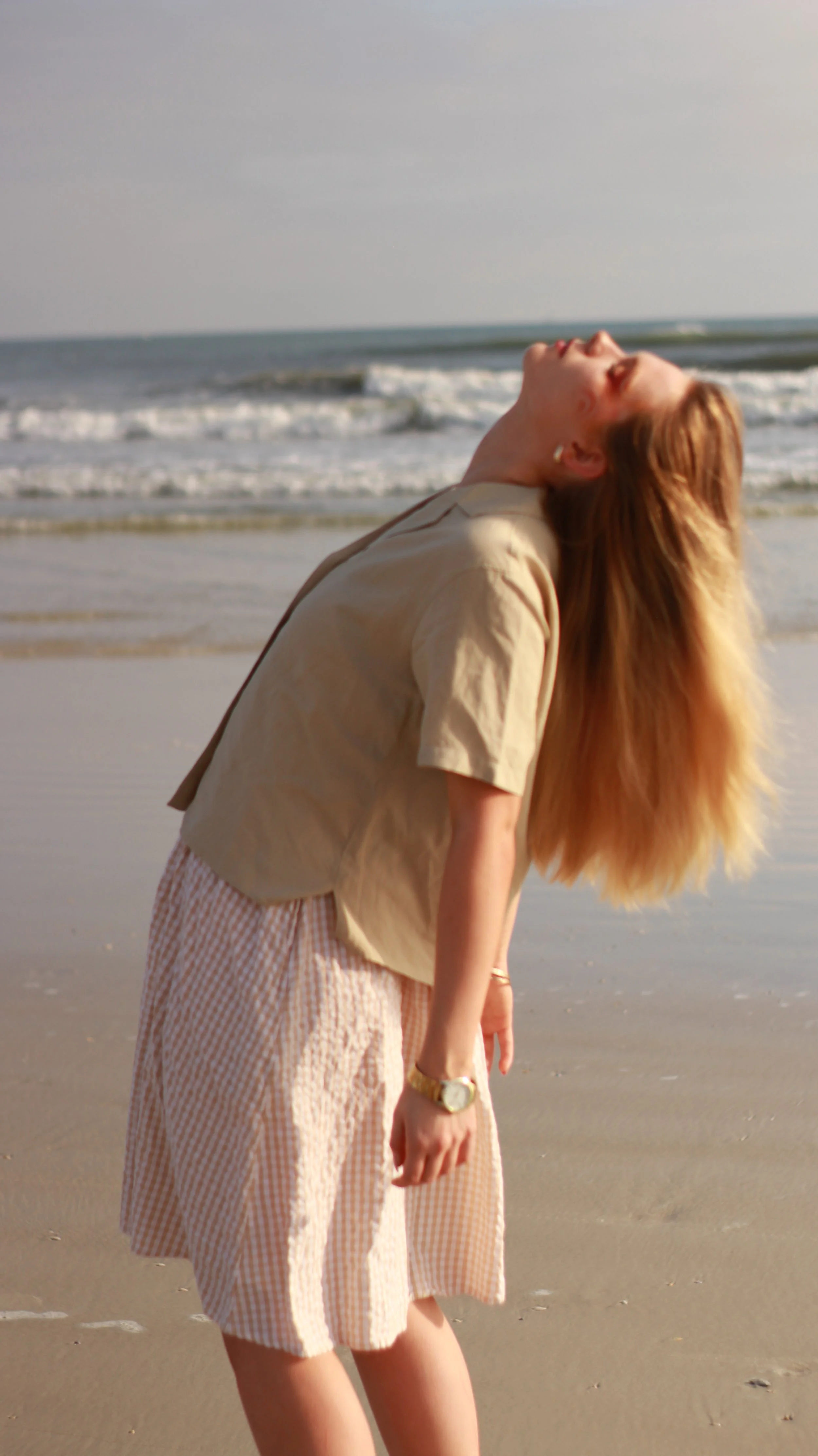 A woman with long red hair wearing a beige jacket and pink checkered pants, standing on a beach with her head tilted back and eyes closed as the ocean waves roll in.