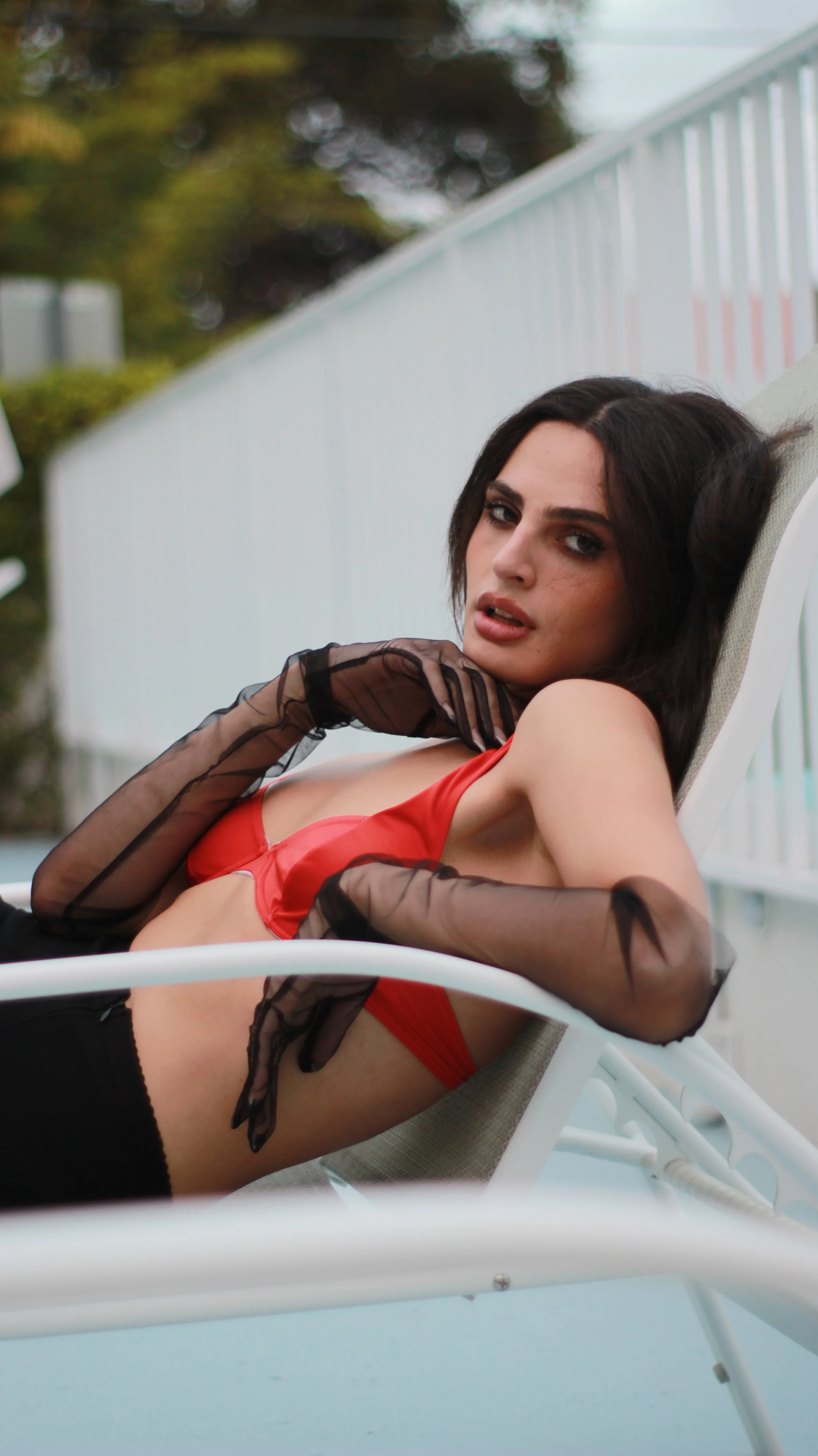 Woman with dark hair wearing red lingerie and black sheer gloves, reclining on pool chair outdoors.