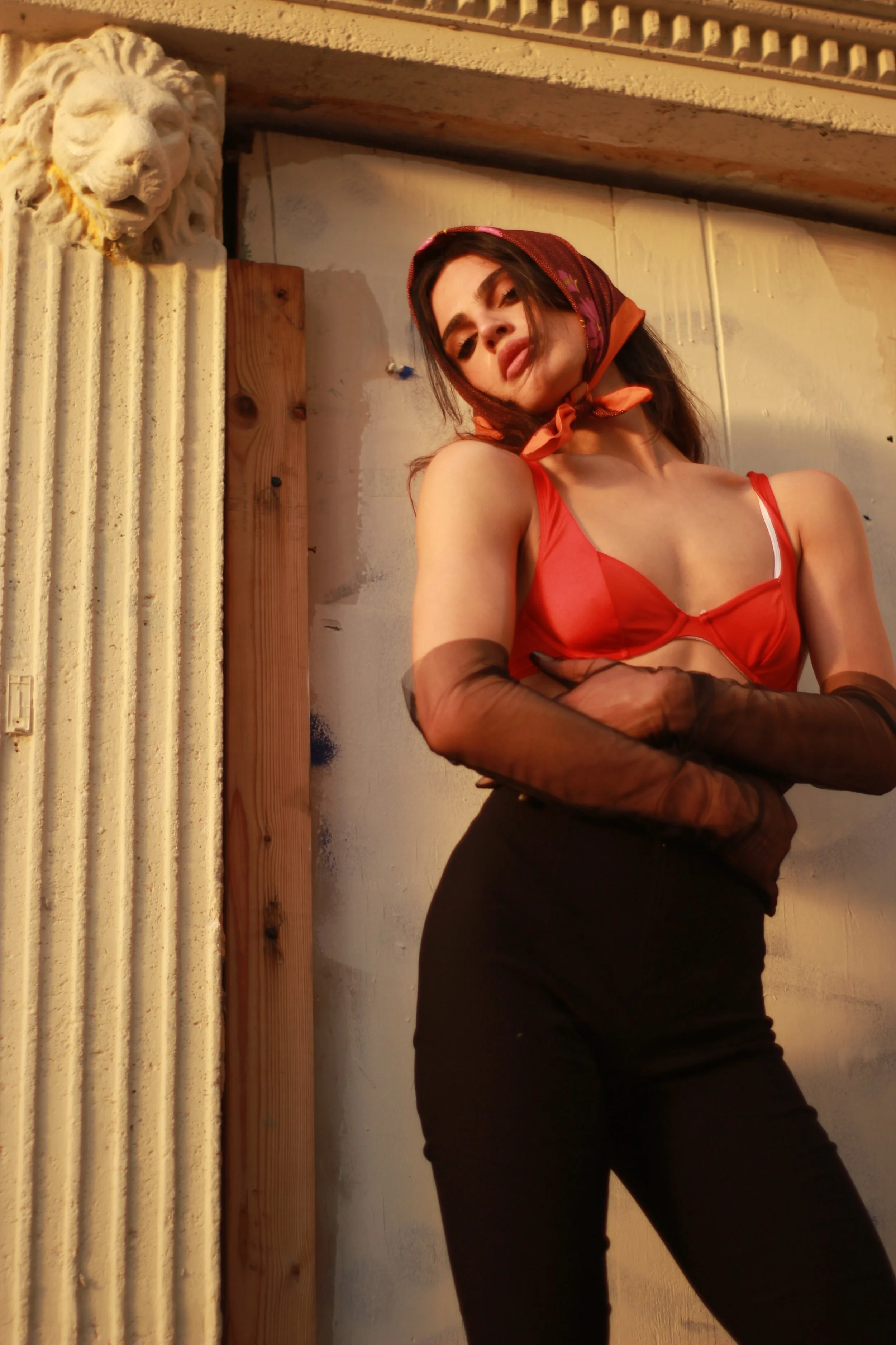 A woman wearing a red bra, black pants, sheer black gloves, and a red headscarf standing against a wall with a lion sculpture on the left side of the image.