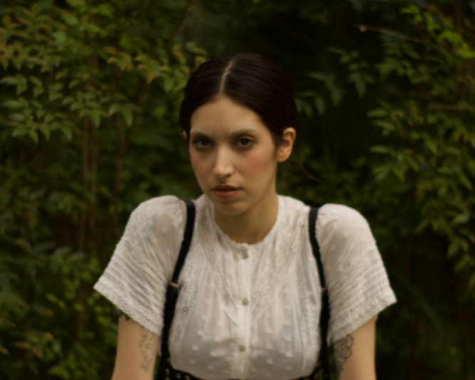 A young woman with dark hair and tattoos wearing a white blouse standing outdoors against green foliage.