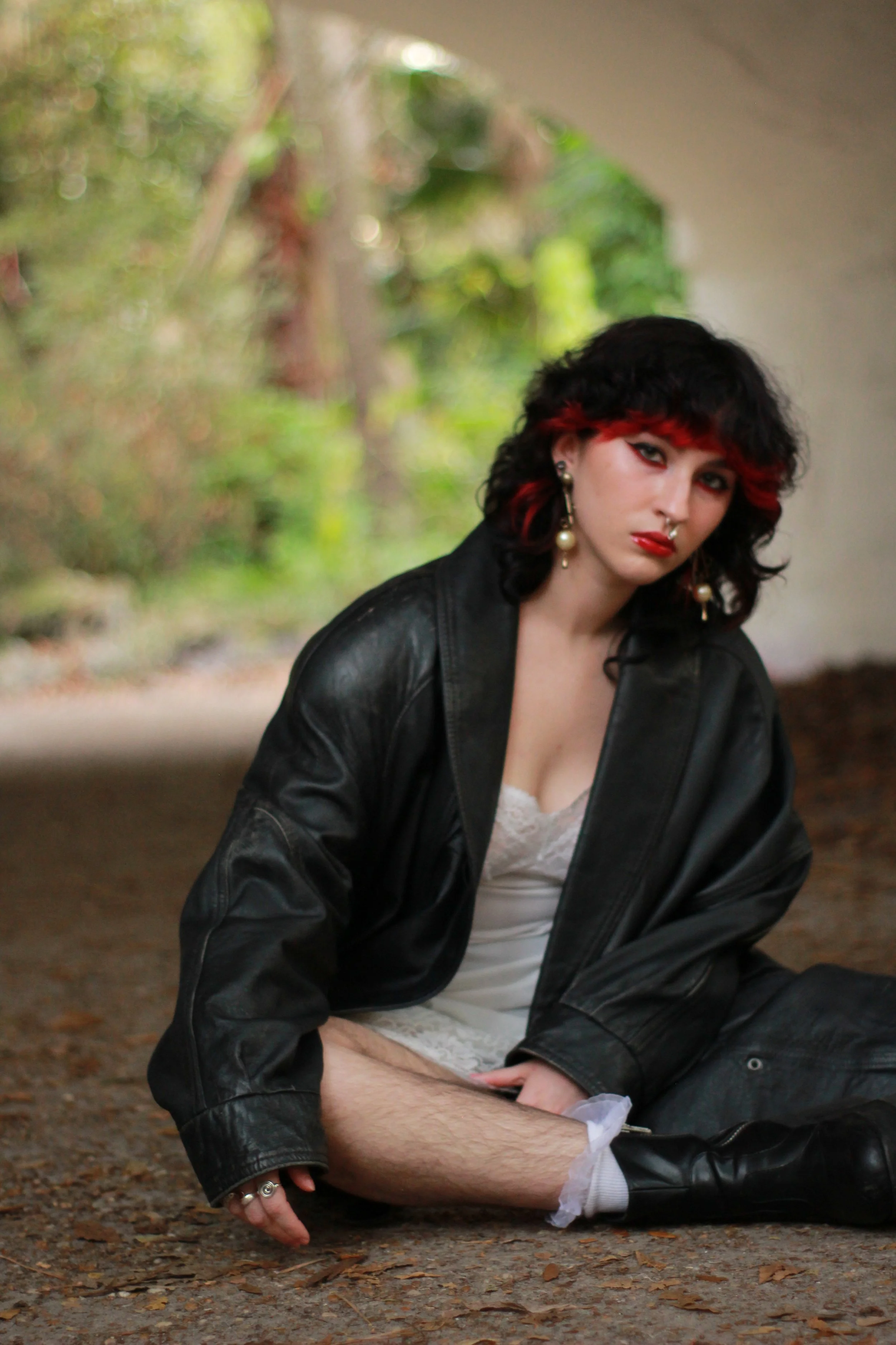 A person with black and red curly hair, makeup, earrings, and a septum piercing sitting on the ground outdoors, wearing a black leather jacket and white lace clothing, with a blurred green natural background.