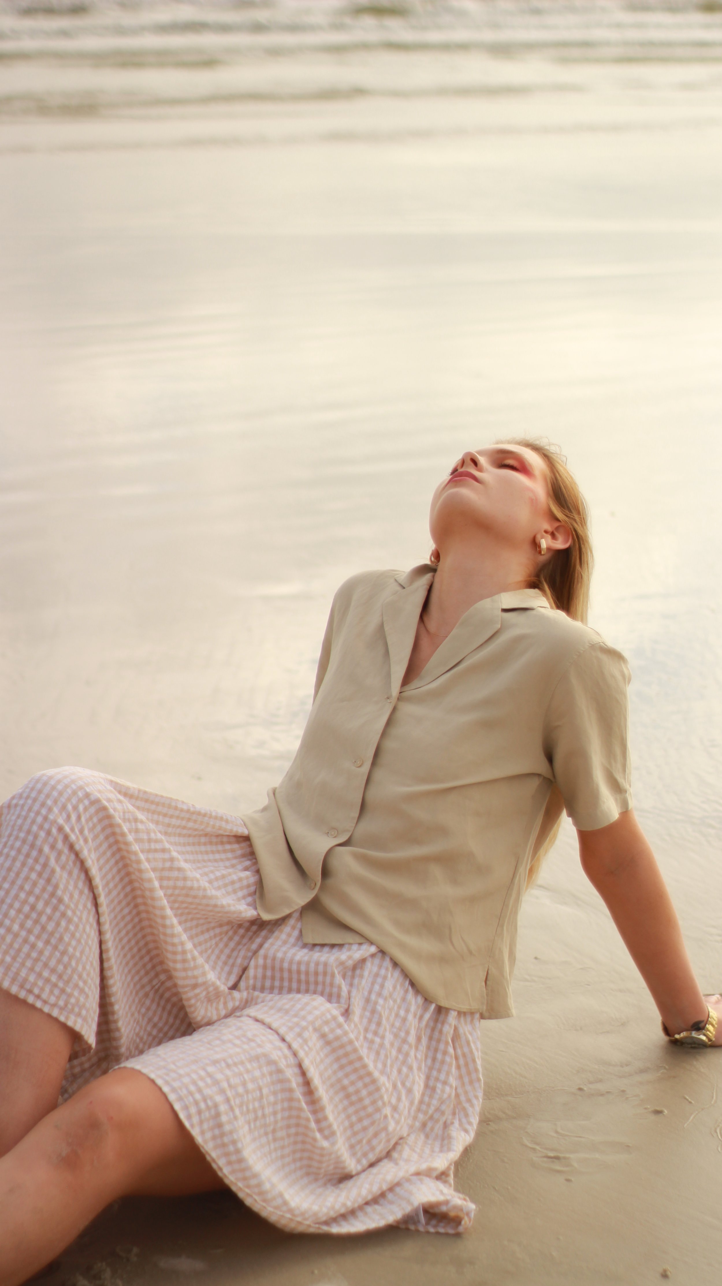 A woman relaxing on a sandy beach with her head tilted back, wearing a beige shirt and checkered shorts, near calm water.