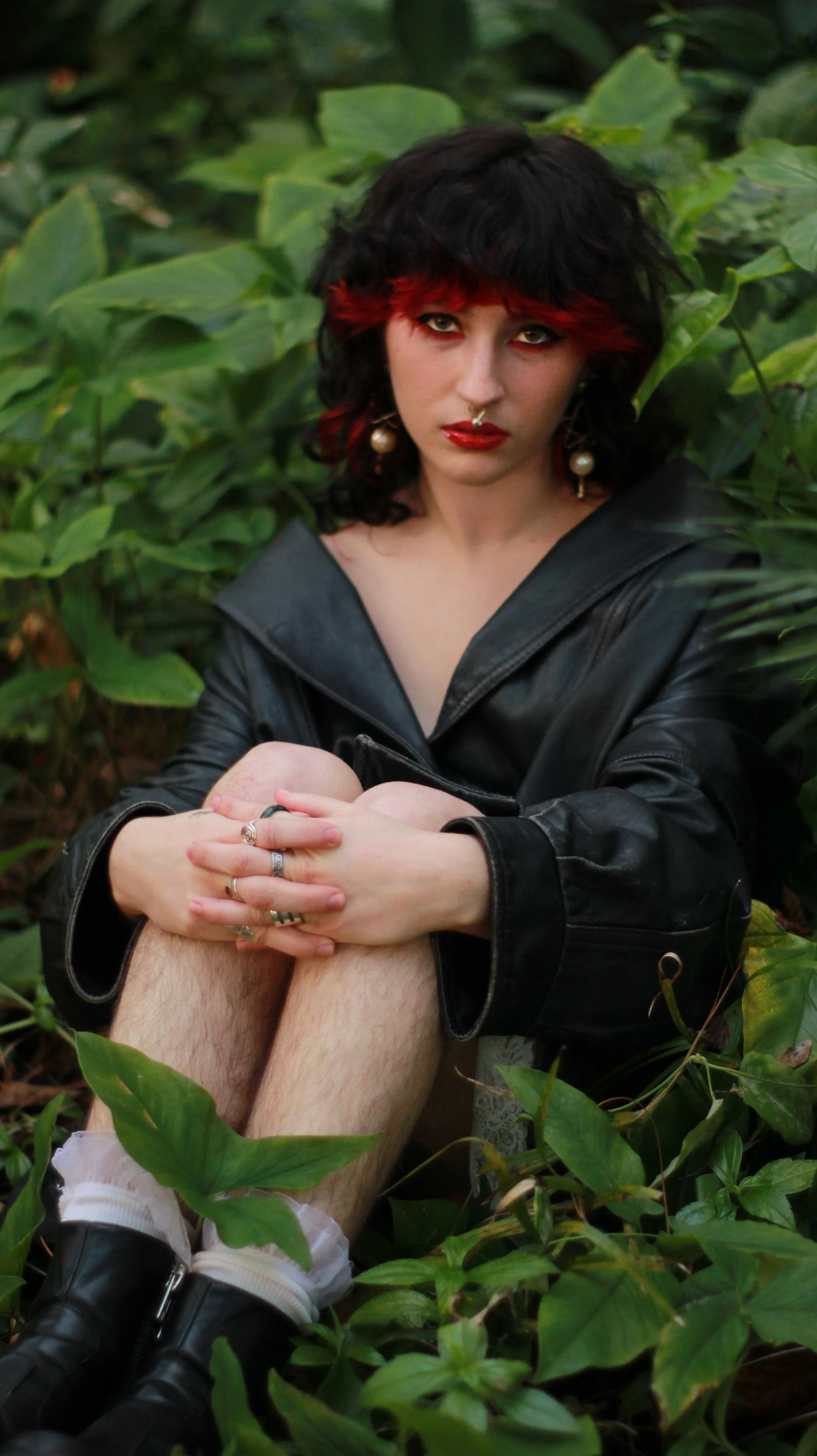 A woman with black and red hair, wearing large pearl earrings and a black leather jacket, sitting among green leafy plants with crossed arms.