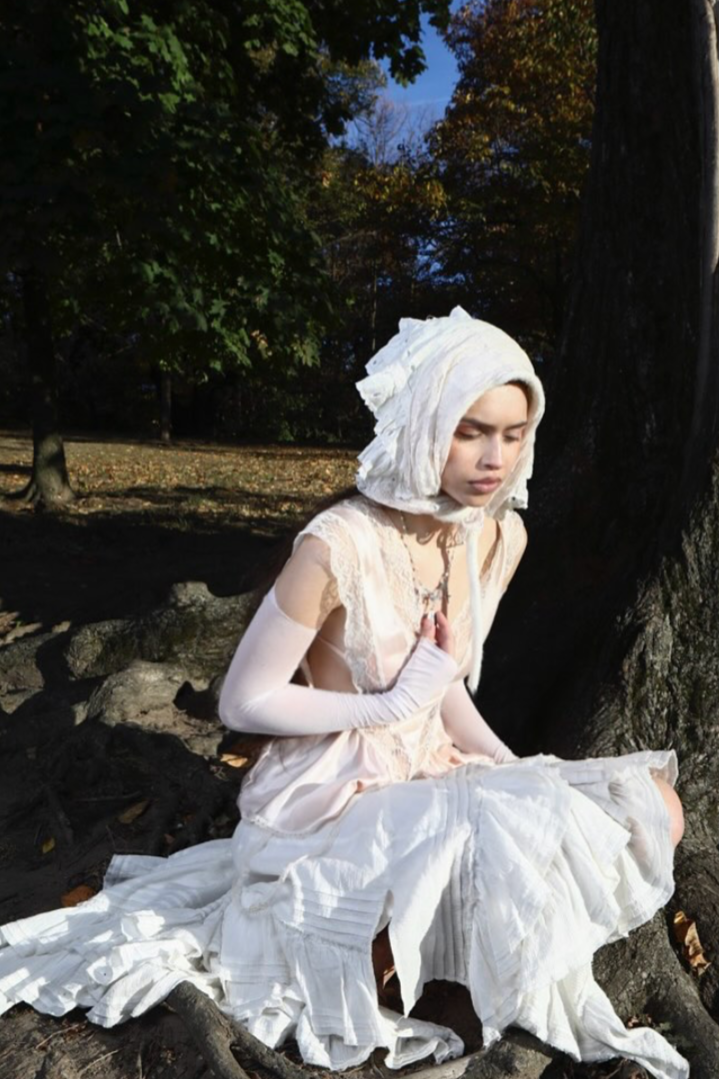 A woman dressed in white layers, sitting outdoors against a large tree in a park-like setting, with her eyes closed and hands in a prayer position.