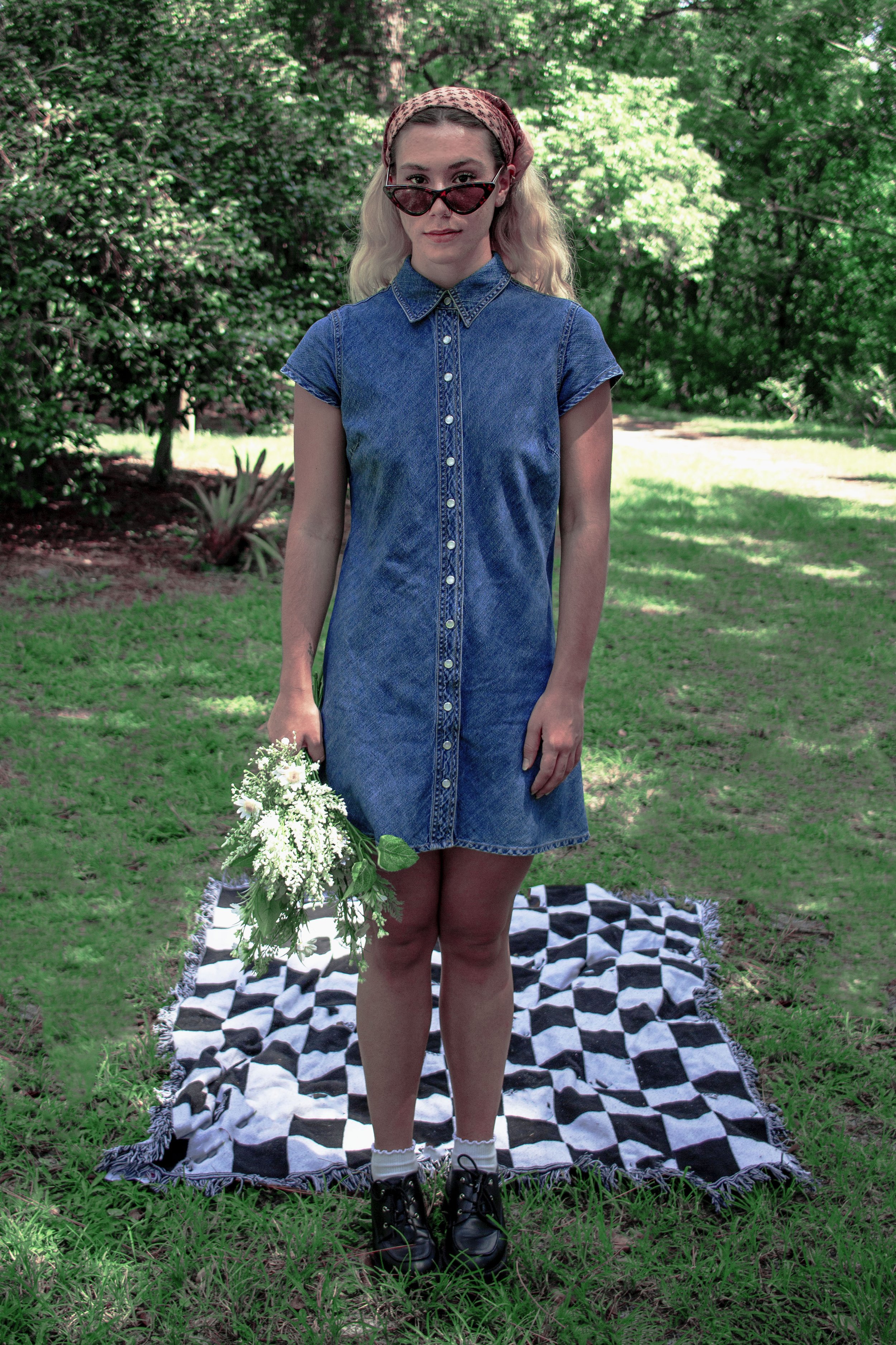 A young woman with blonde wavy hair, wearing a red headscarf, dark sunglasses, a sleeveless denim dress with small white buttons, and black combat boots, stands on a black and white checkered blanket in a park with lush green trees, holding a bouquet