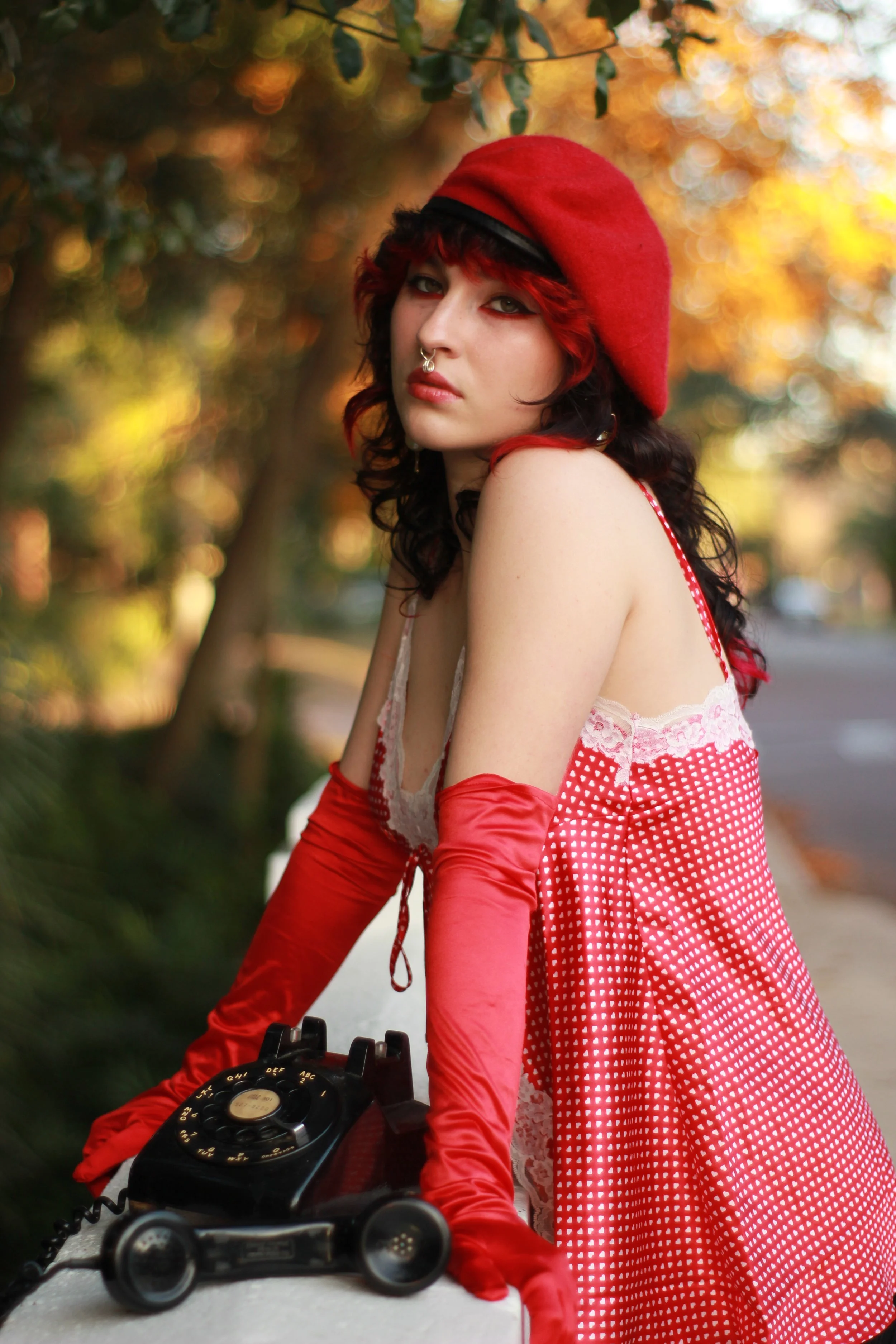 A young woman with dark curly hair and light skin is wearing a red beret, red gloves, and a pink and red polka dot dress. She is sitting outdoors, with blurred autumn trees in the background, and looking at the camera with a serious expression while 