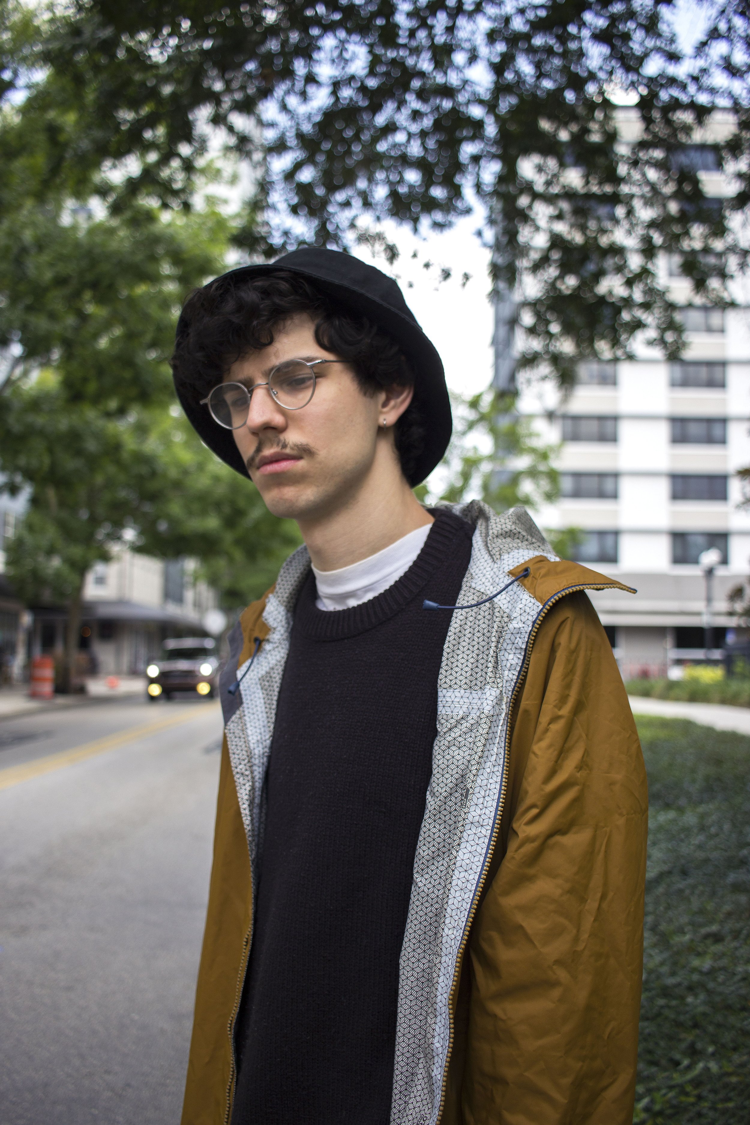 A young man with curly dark hair, wearing glasses, a black hat, a black sweater, and a brown jacket, standing outdoors in a city park during daytime, with trees and a tall building in the background.