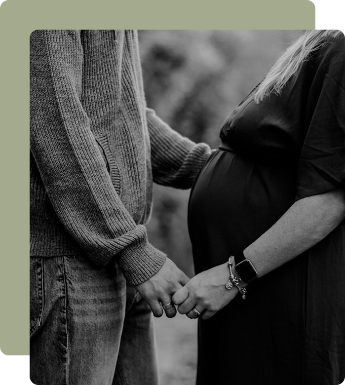 A pregnant woman and her partner holding hands outdoors, with the woman's pregnant belly visible, in black and white.