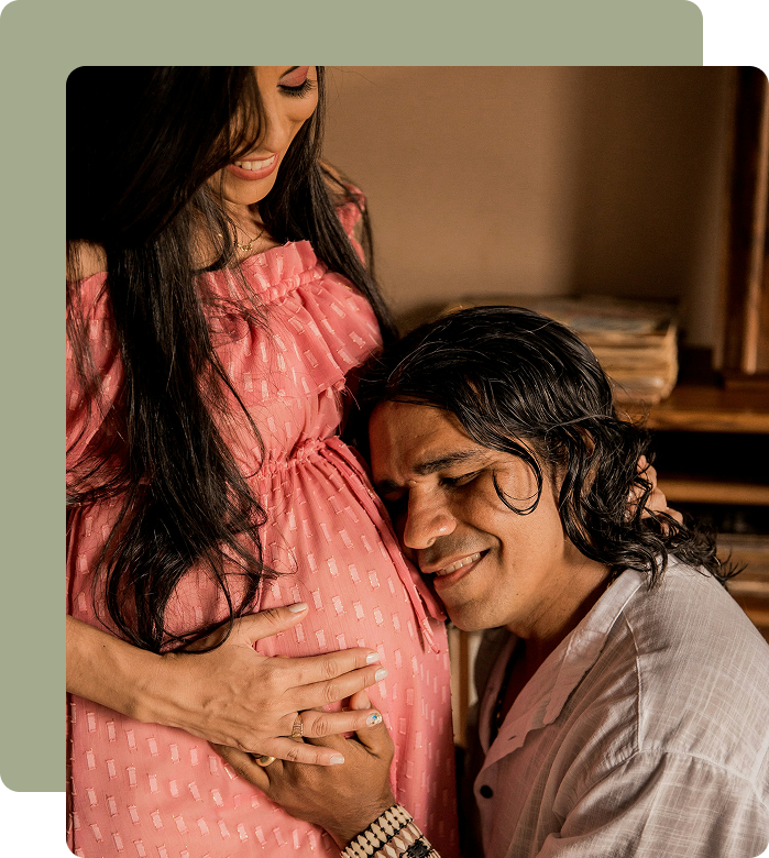A man kneeling with his head on a pregnant woman's belly, gently touching it. The woman, wearing a pink dress, is smiling and holding her belly with one hand. The scene is warm and intimate, indicating a moment of connection and anticipation for a baby.