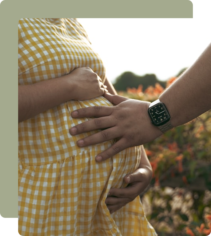 Person in yellow and white checkered dress holding their pregnant belly with one hand, while another person touches the belly with their hand, wearing a smartwatch. Background features outdoor plants and sunlight.