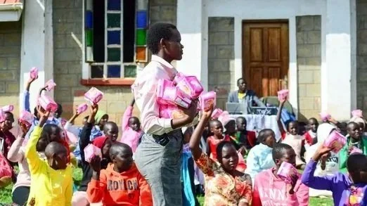 A woman stands in front of a crowd of children, holding several pink packages, as children raise similar packages in the air outdoors near a building.
