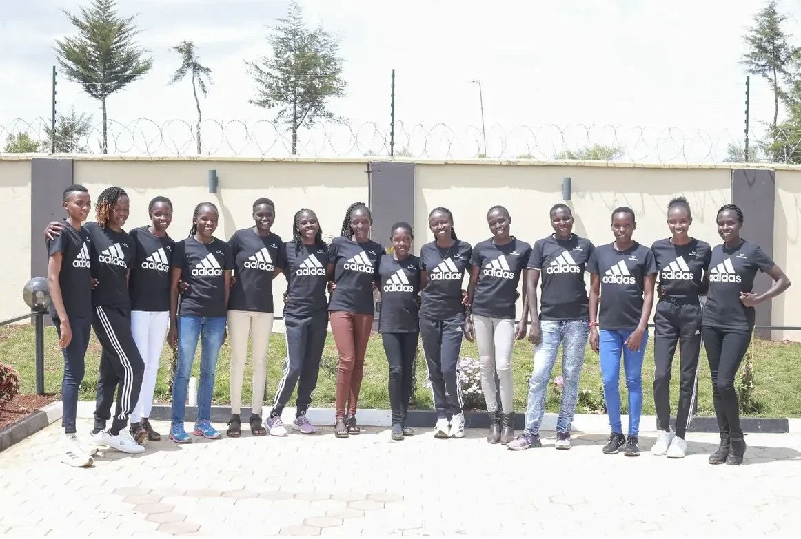 A group of 14 young women standing outdoors in front of a wall with barbed wire, all wearing black Adidas shirts and jeans or pants, smiling for the photo.