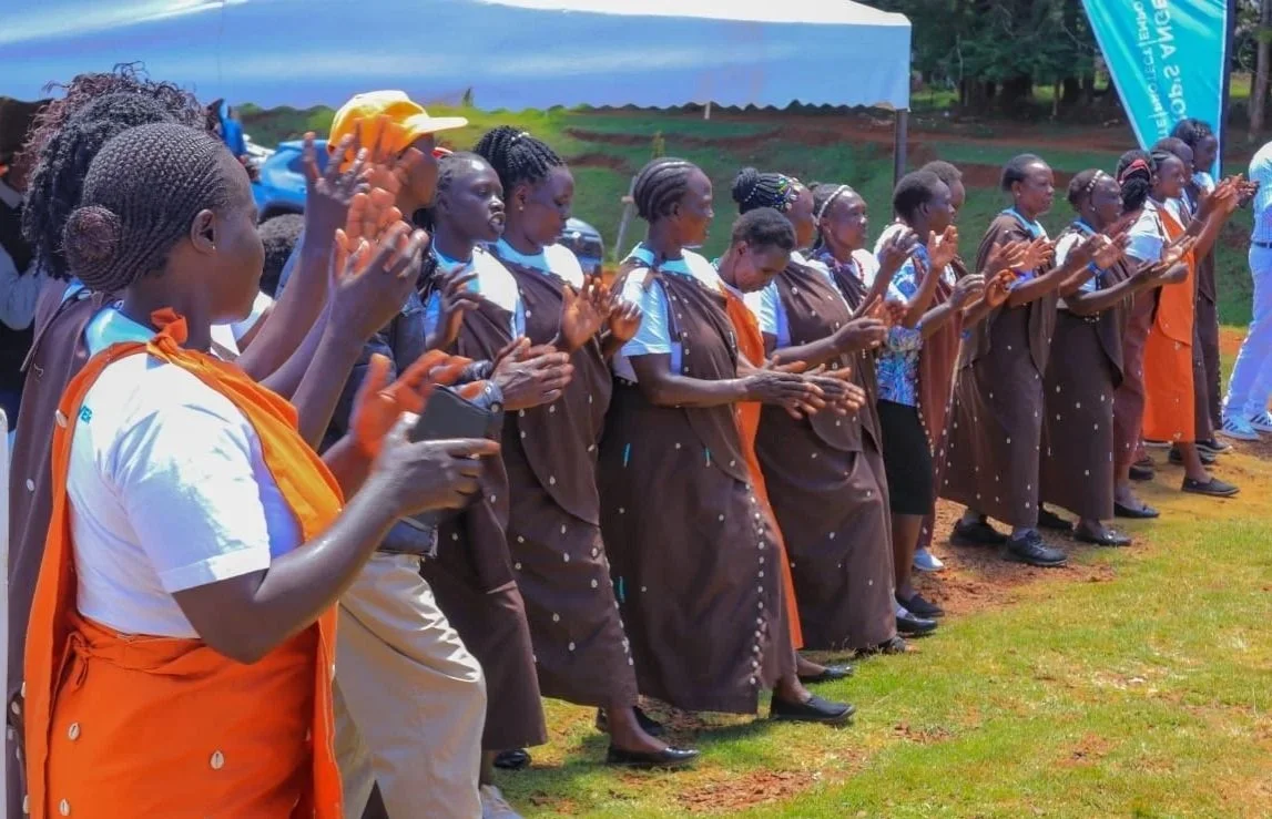 Group of women with braided hair, dressed in brown dresses with orange accents, participating in an outdoor event, clapping and singing under a blue tent.