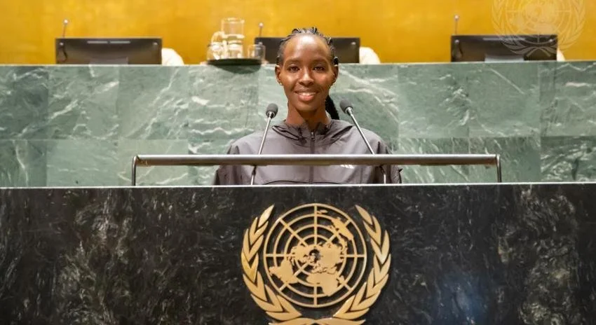 A woman in a black robe standing at a UN podium, smiling, with microphones in front of her, in a room with green marble walls and the UN emblem.