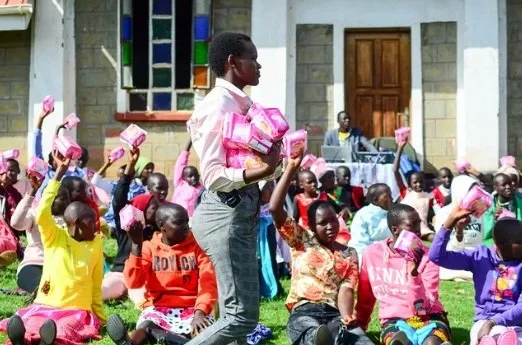 A group of children and a woman sitting and standing in front of a building, holding pink boxes, during an outdoor event at a school or community center.