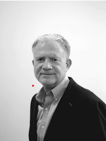 Black and white photo of a man with short, light-colored hair, wearing a collared shirt and blazer, standing against a plain background.