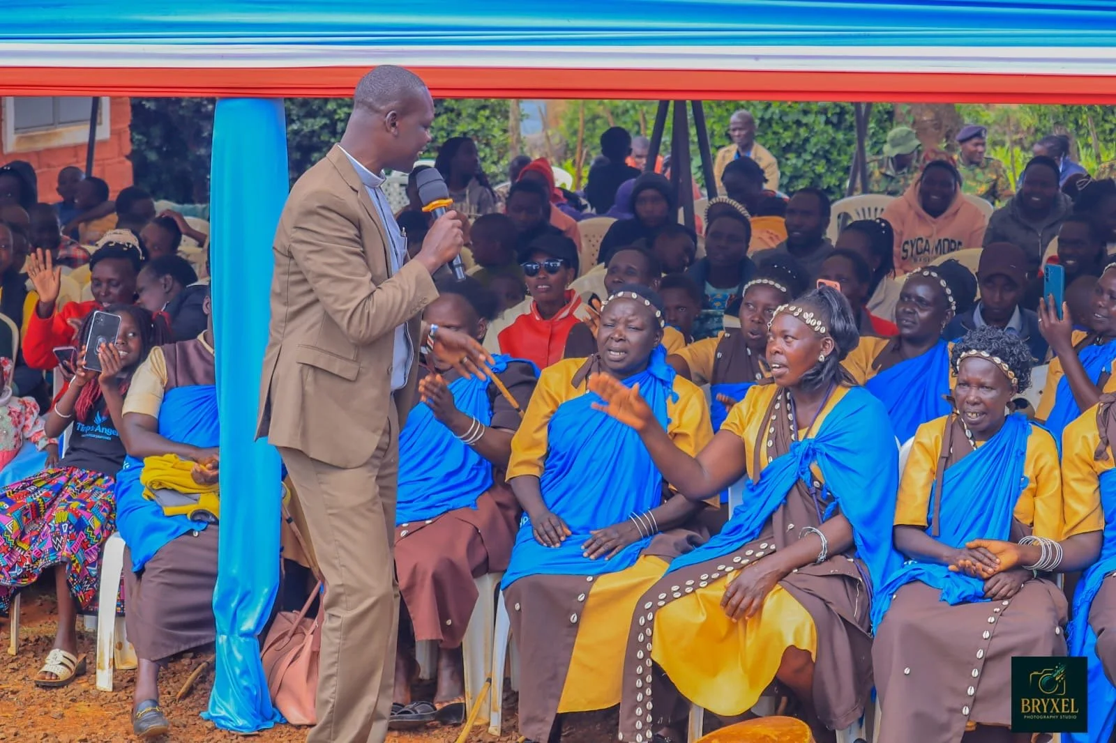 Man in beige suit holding microphone speaking to a group of women dressed in blue and yellow, seated outdoors under a tent during daytime, with many people in the background.