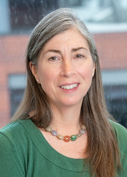 Portrait of a middle-aged woman with gray hair, smiling, wearing a green top and a colorful beaded necklace, standing outdoors in front of a blurred background.
