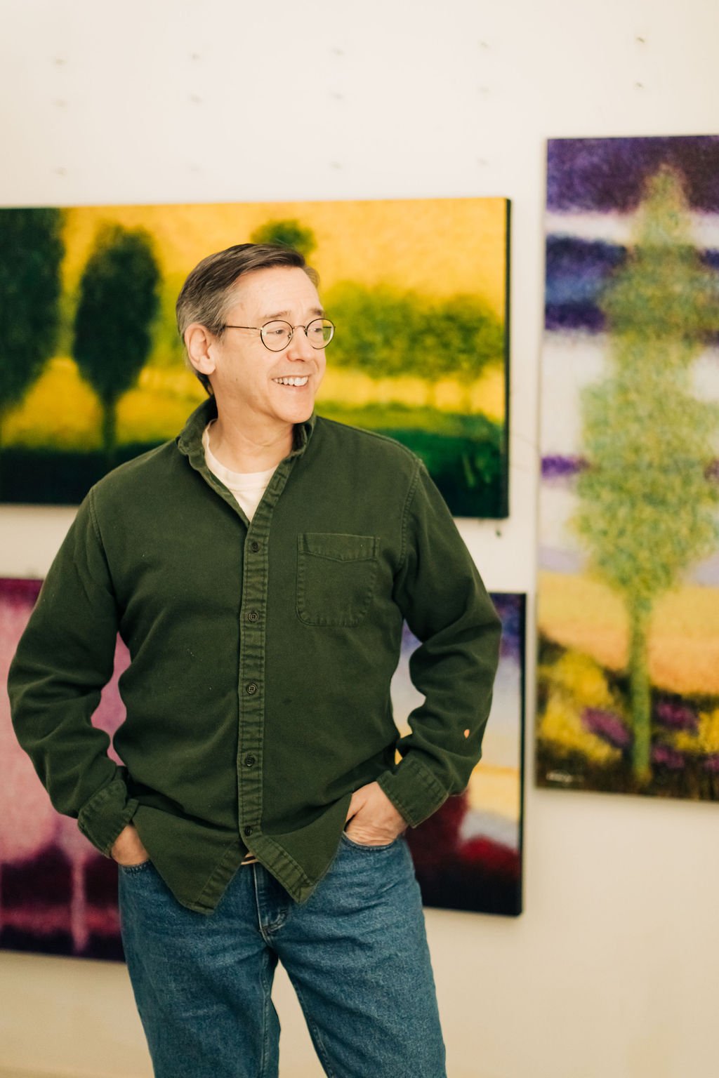 A man with glasses and gray hair smiling at an art gallery, standing in front of colorful landscape paintings.