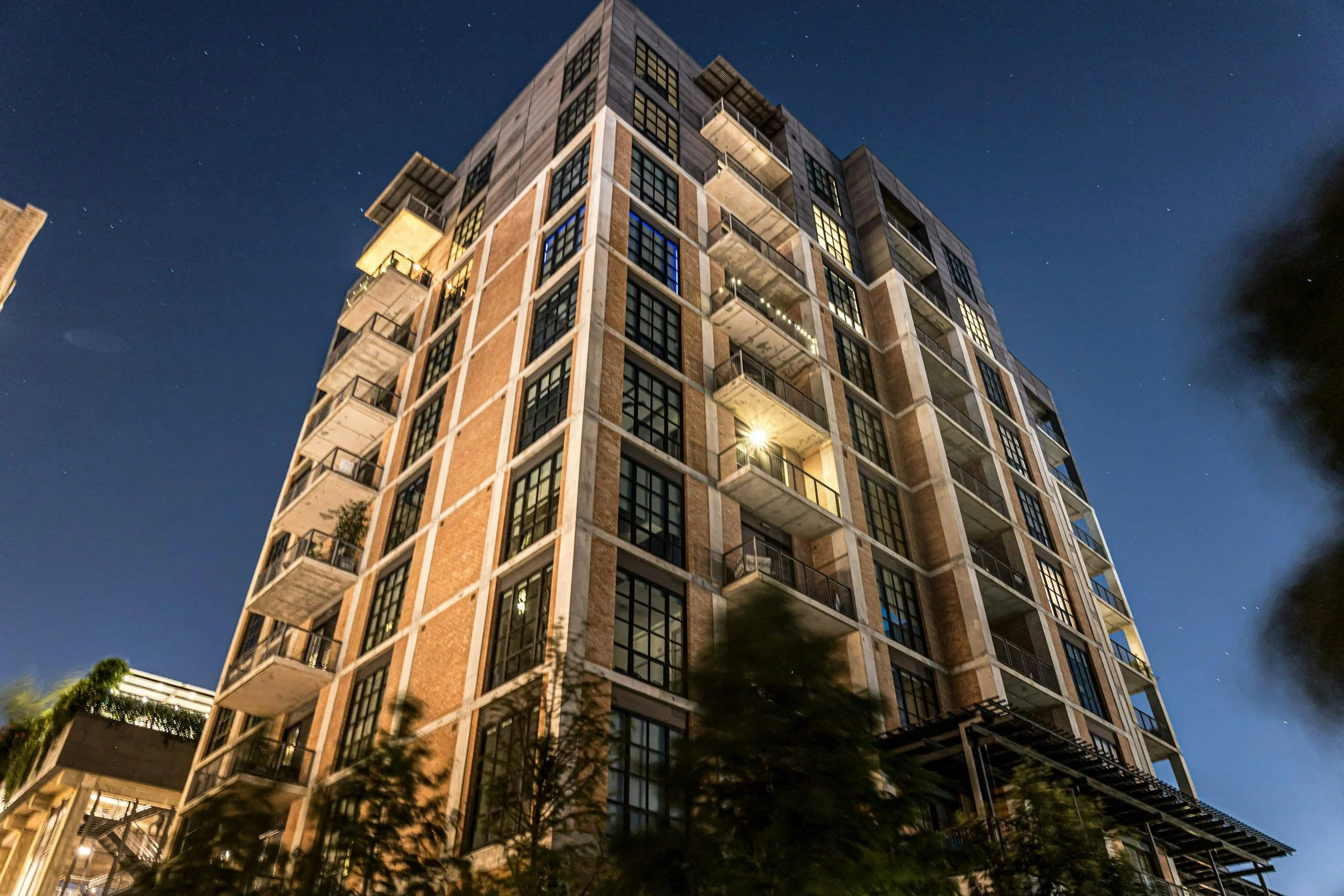 Nighttime view of a modern multi-story residential building with large windows and balconies, illuminated from within, against a dark sky with stars.