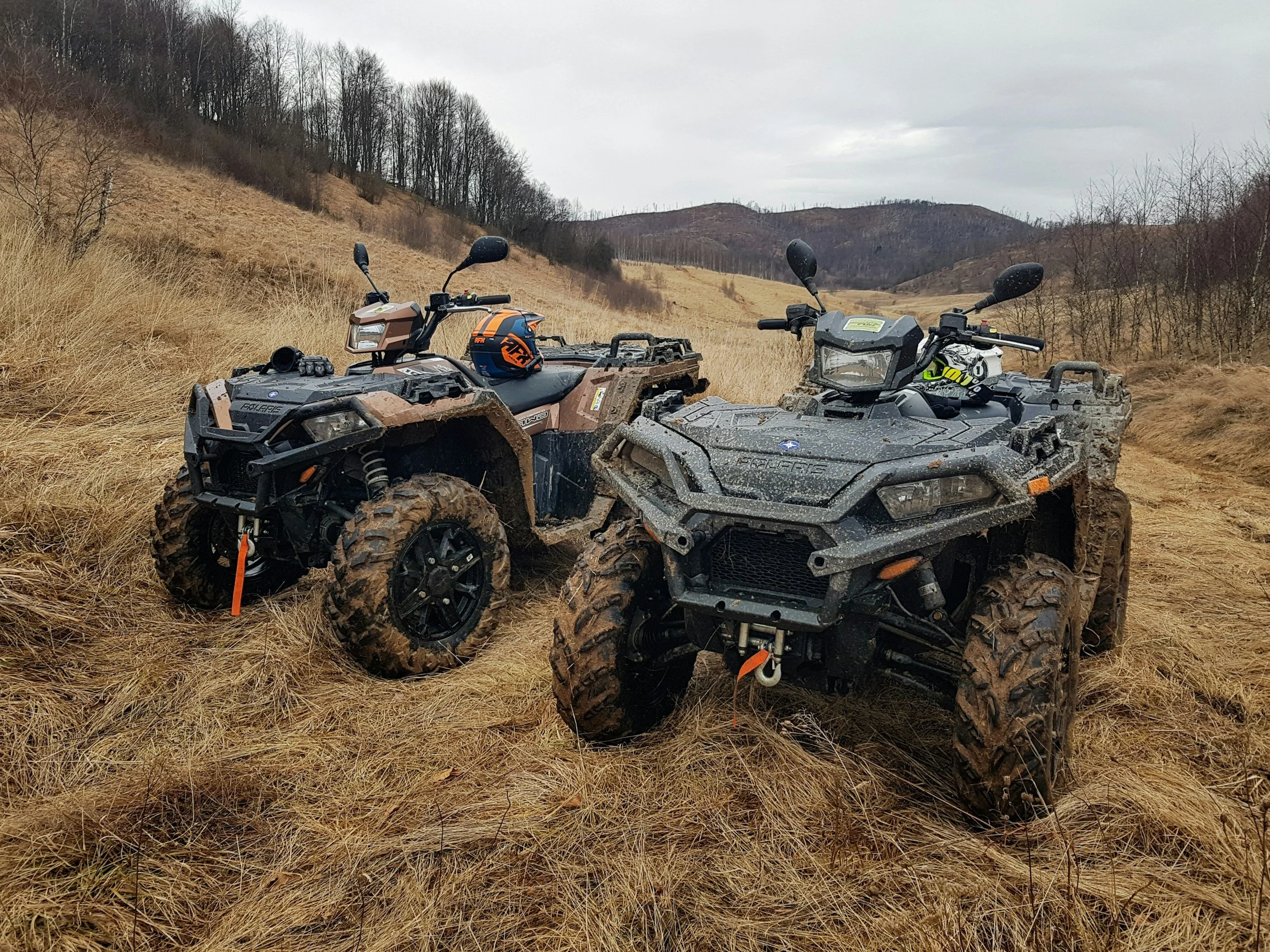 Two all-terrain vehicles (ATVs) parked on a grassy hill with leafless trees and distant hills in the background, under a cloudy sky.
