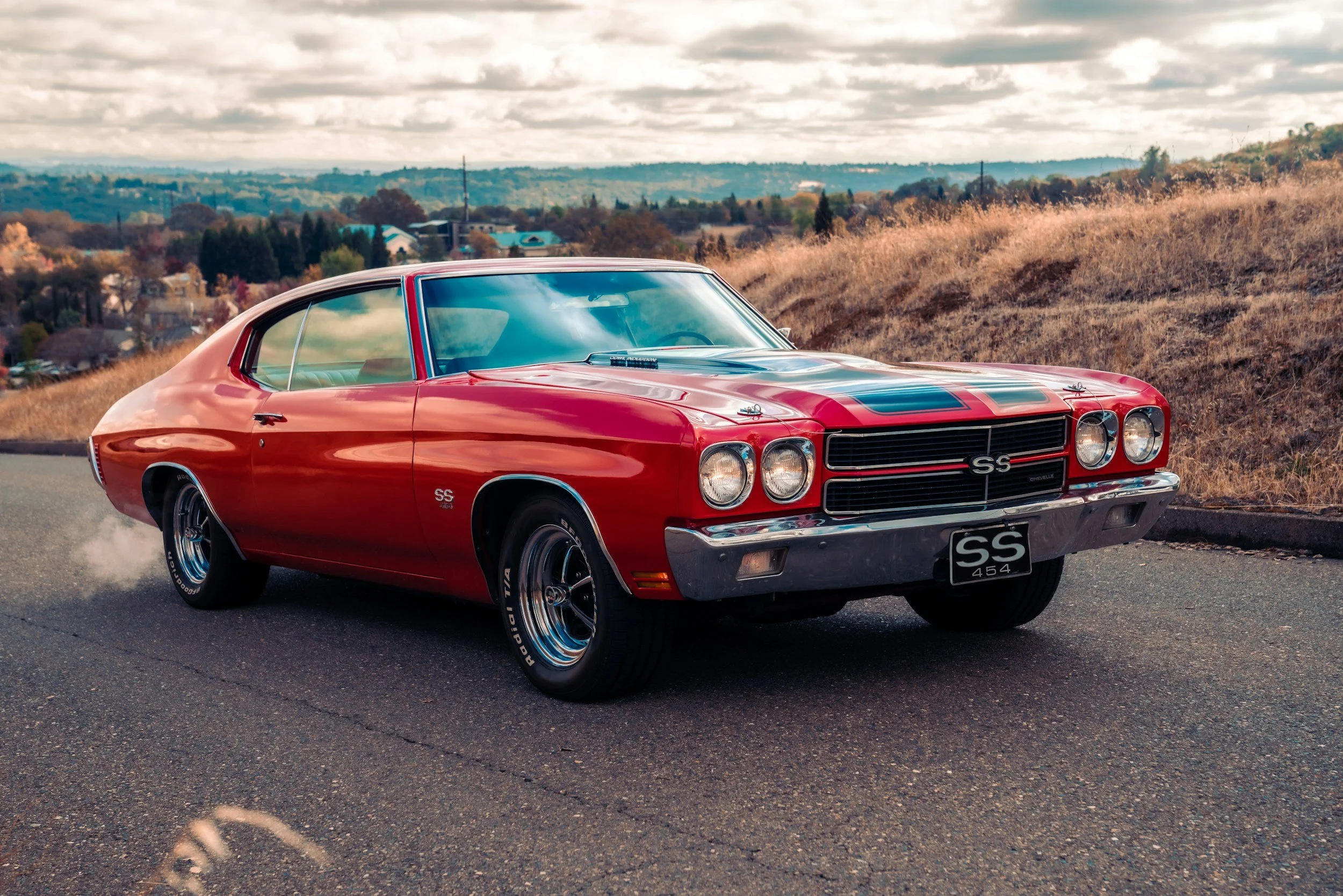 Red vintage Chevrolet SS muscle car driving on a rural road with a scenic landscape of hills and trees in the background.