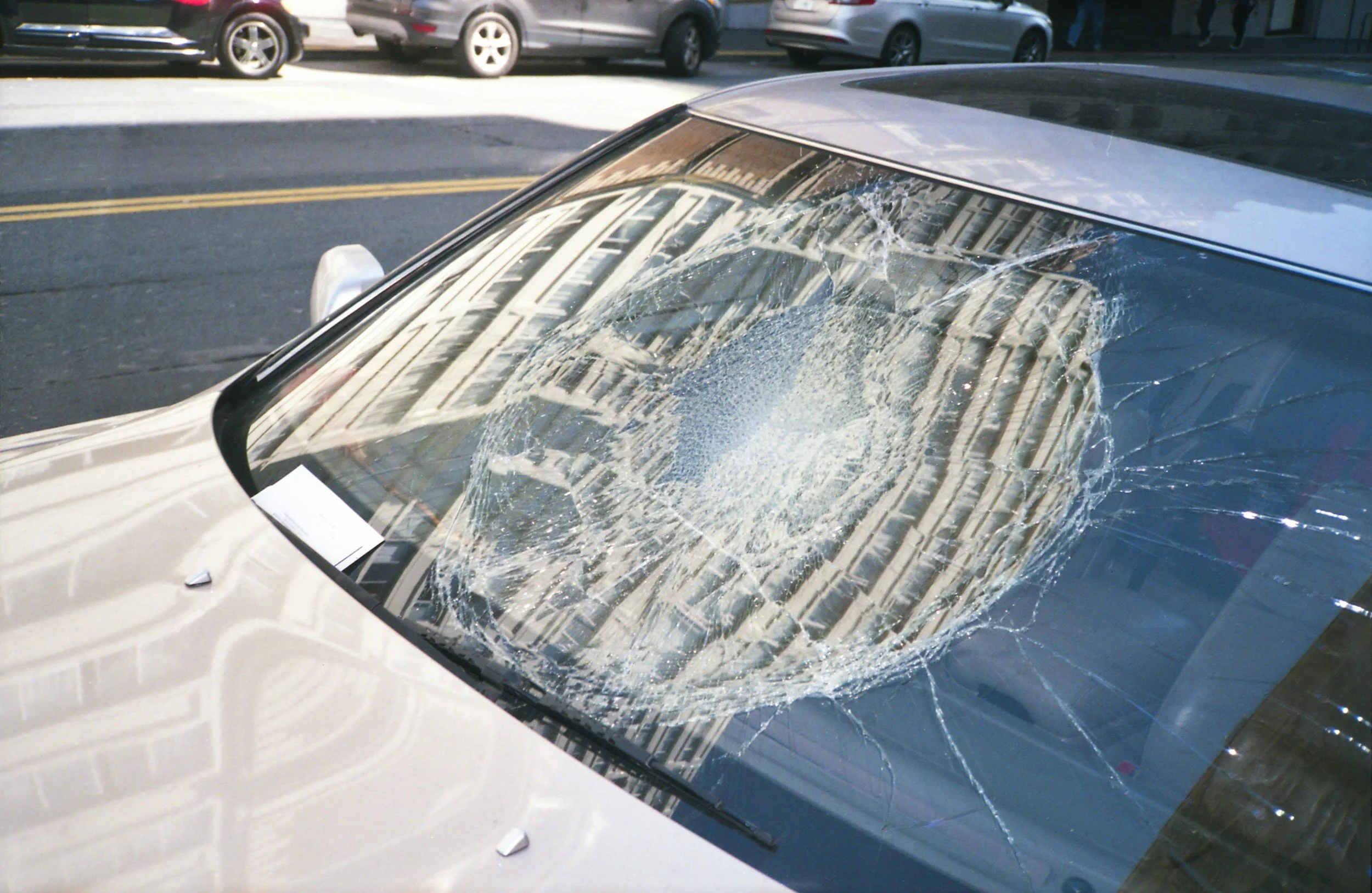A silver SUV with a severely cracked windshield parked on the street, with other parked cars and buildings in the background.